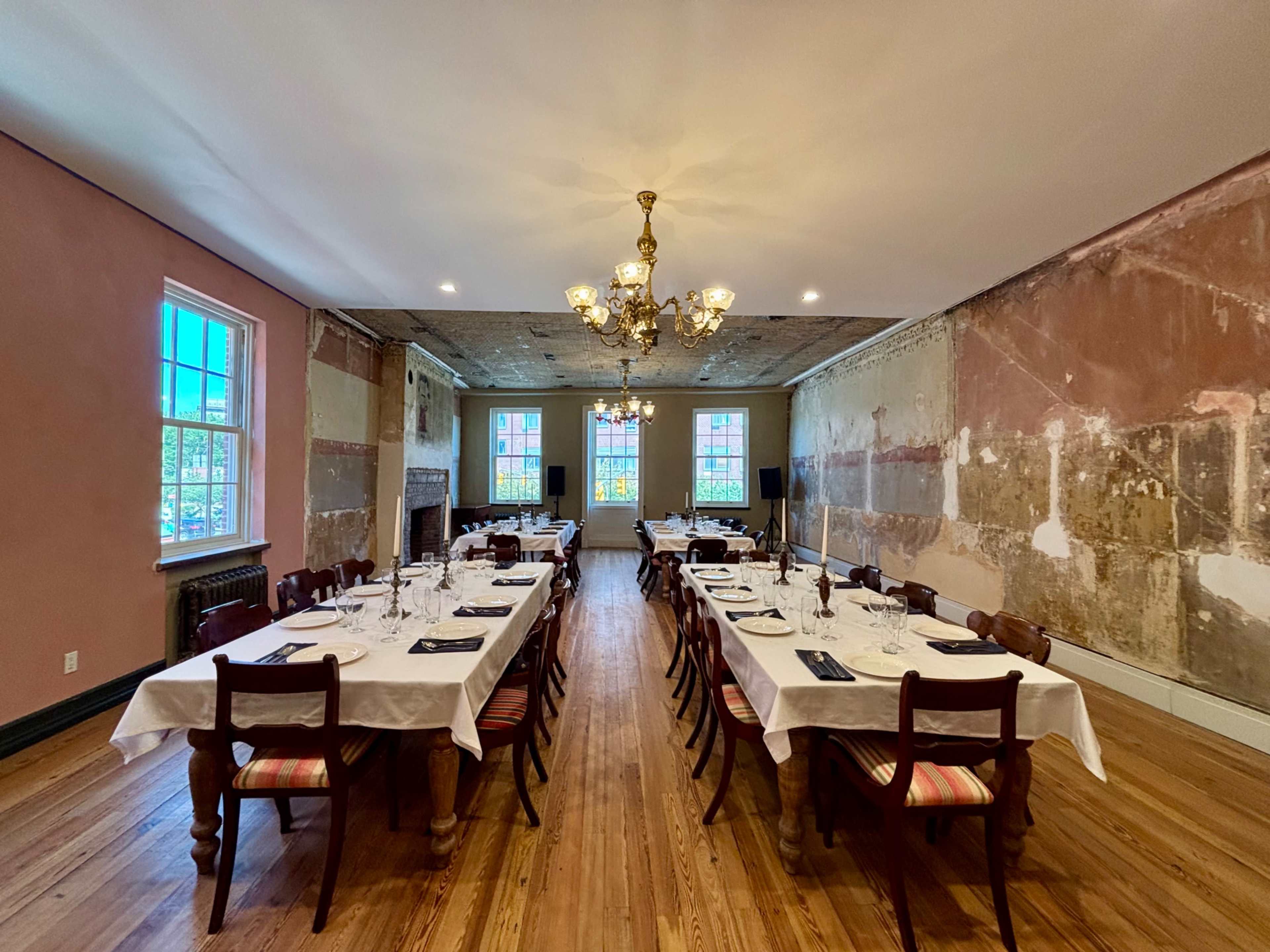 The image shows a dining room with several long tables set with white tablecloths and dining ware, surrounded by walls that have a partially restored, textured finish.