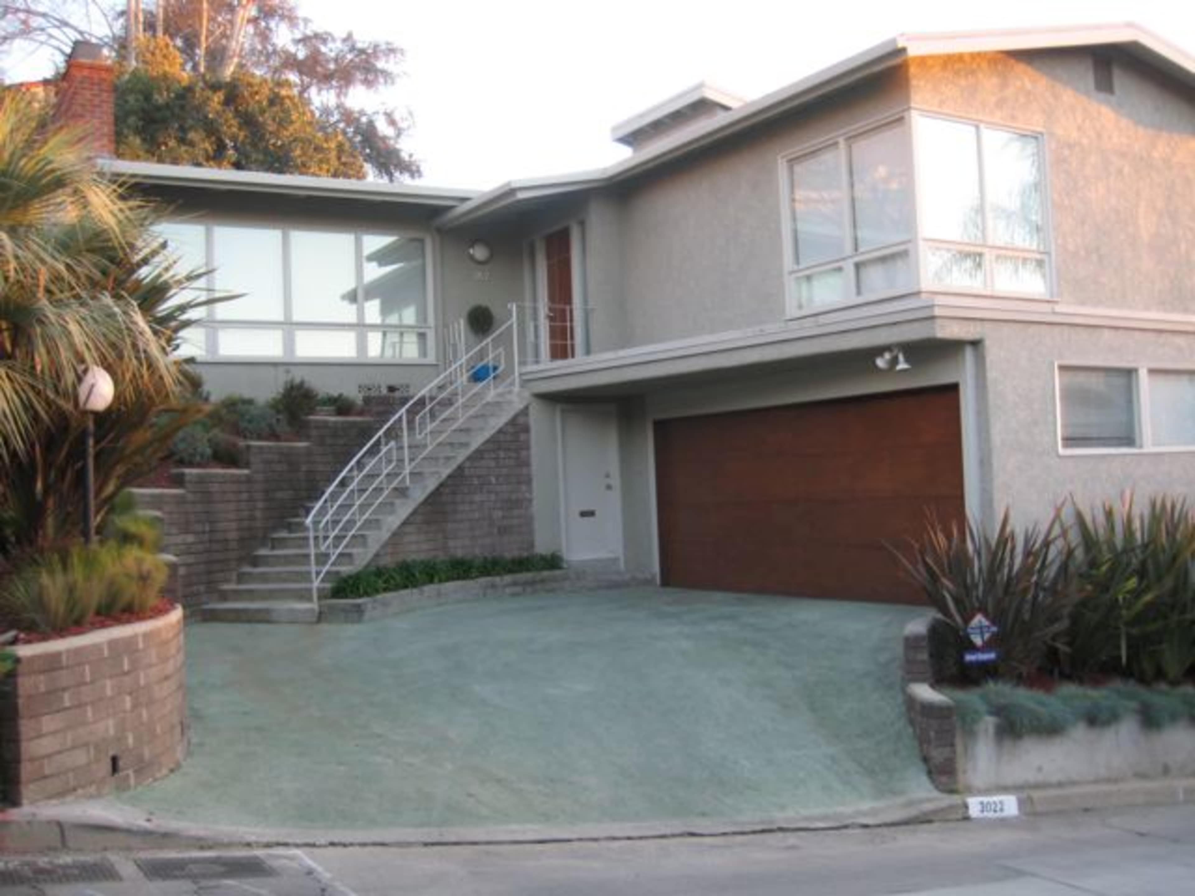 The image shows a modern two-story house with a sloped driveway and a landscaped front yard.