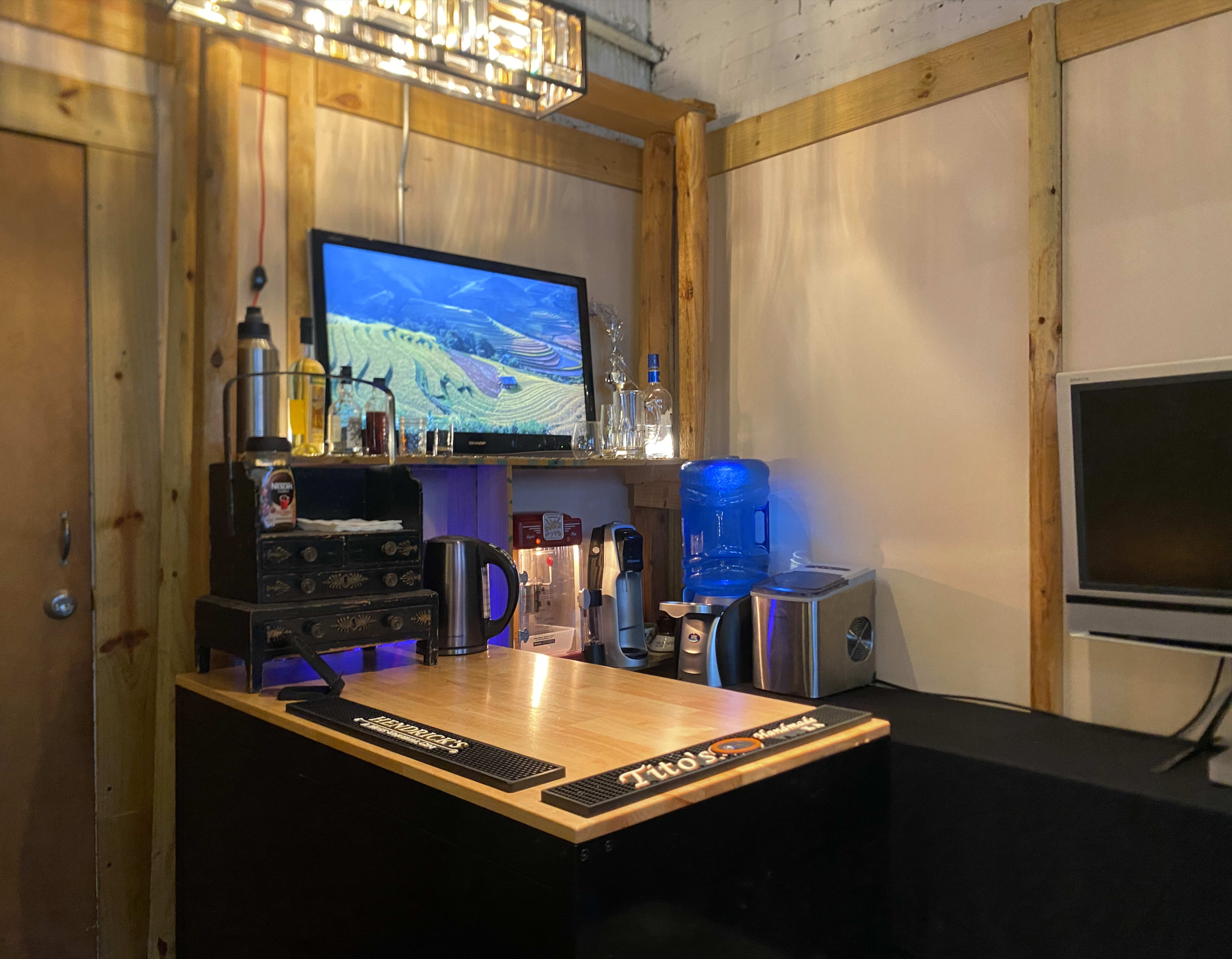 A bar area equipped with a wooden countertop, a television displaying a landscape, bottles of alcohol, a coffee maker, and a water cooler.