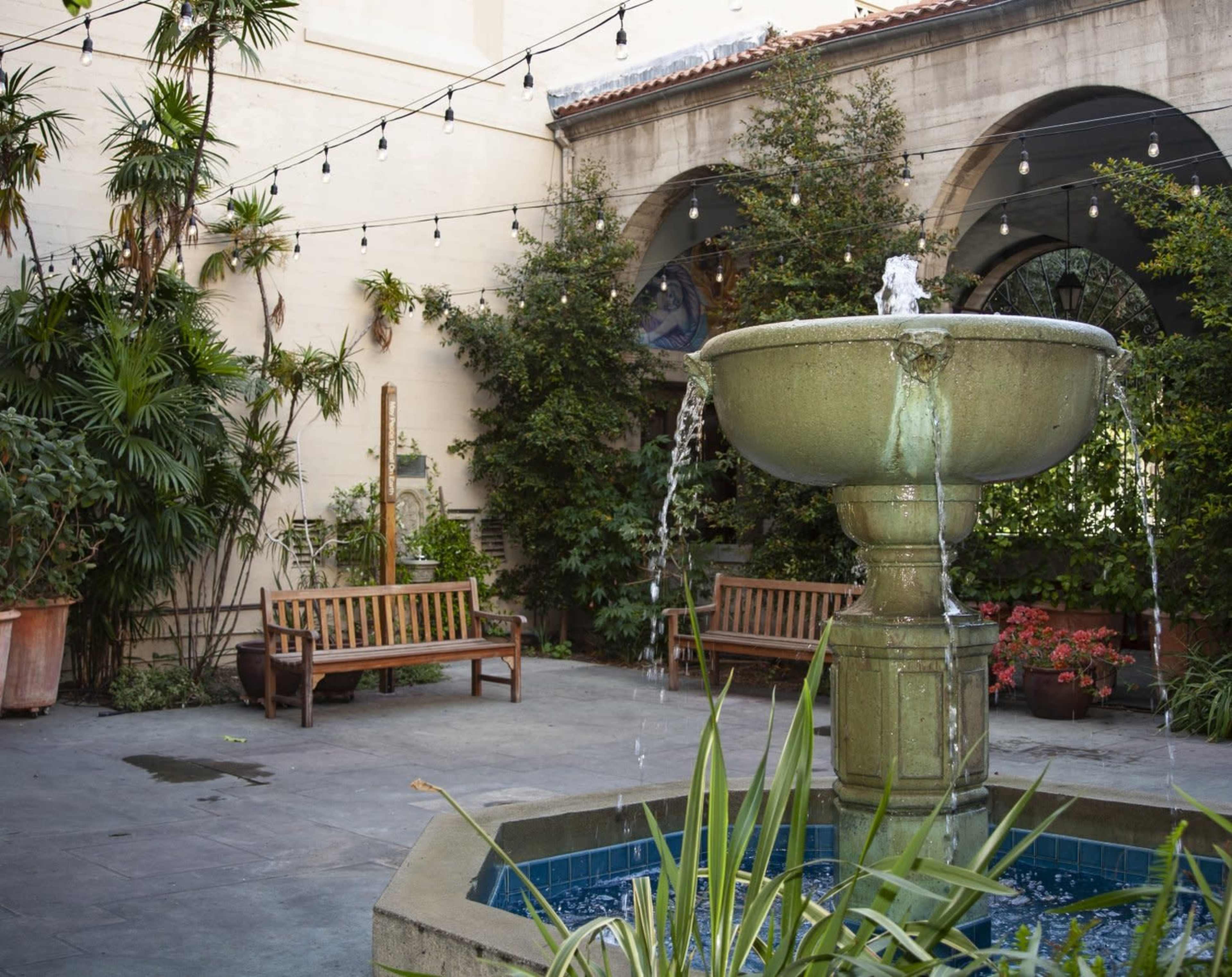 A stone fountain with flowing water is situated in a courtyard surrounded by plants and benches.