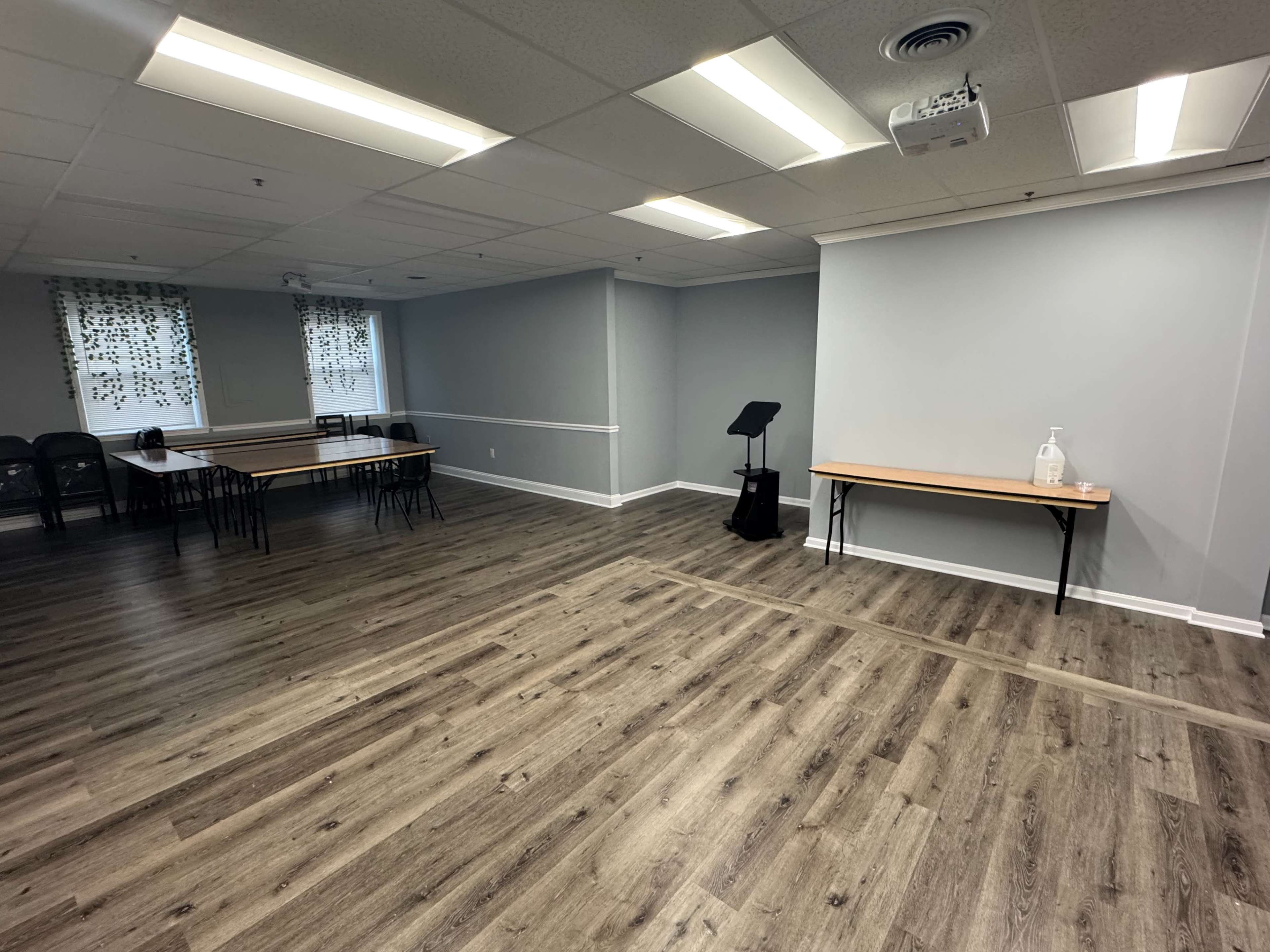 The image shows a sparsely furnished room with several tables and chairs arranged on a wooden floor, and a projection screen mounted on one wall.