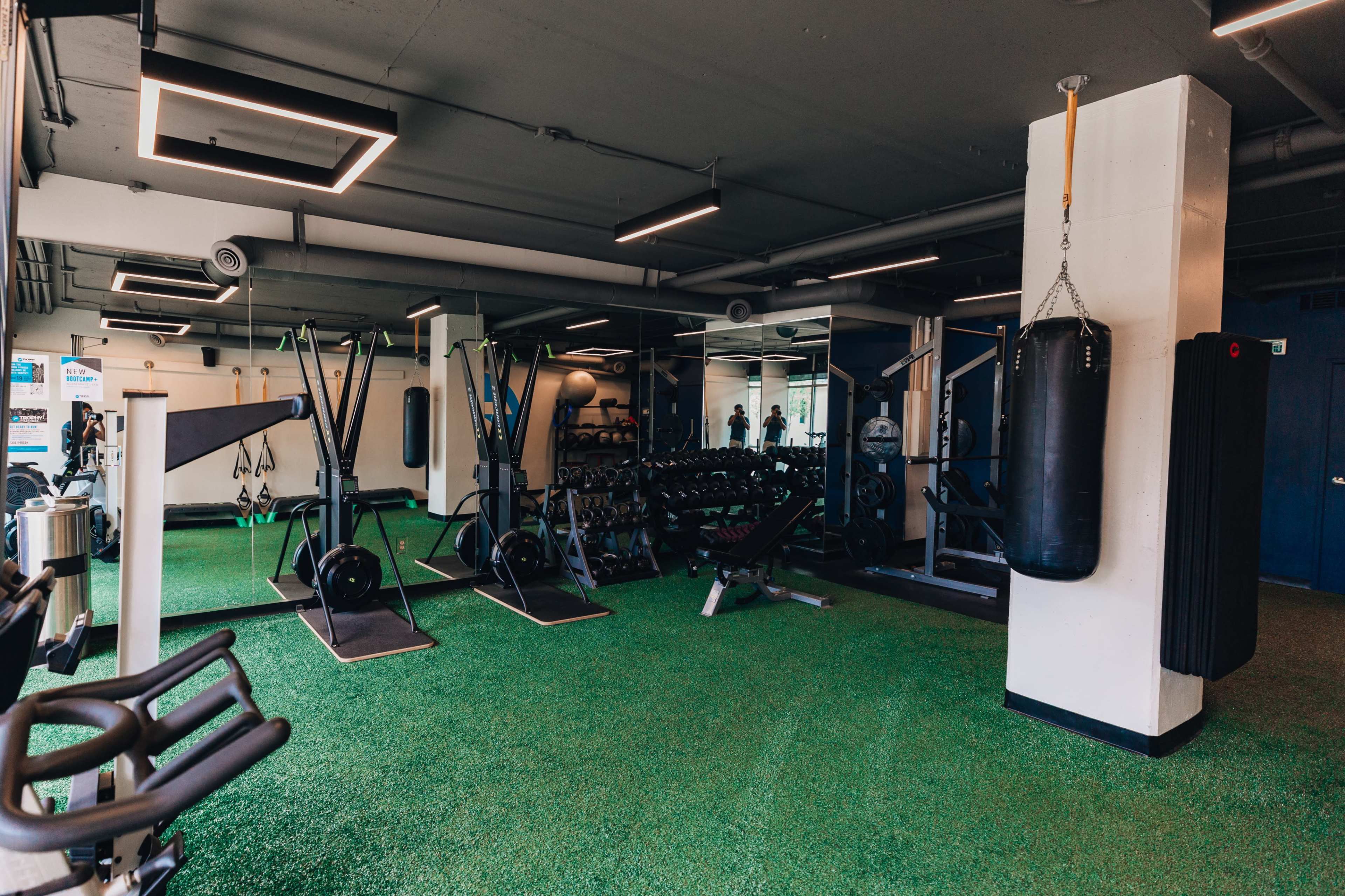 The image shows a fitness gym equipped with various exercise machines, free weights, and a punching bag on a green turf floor.