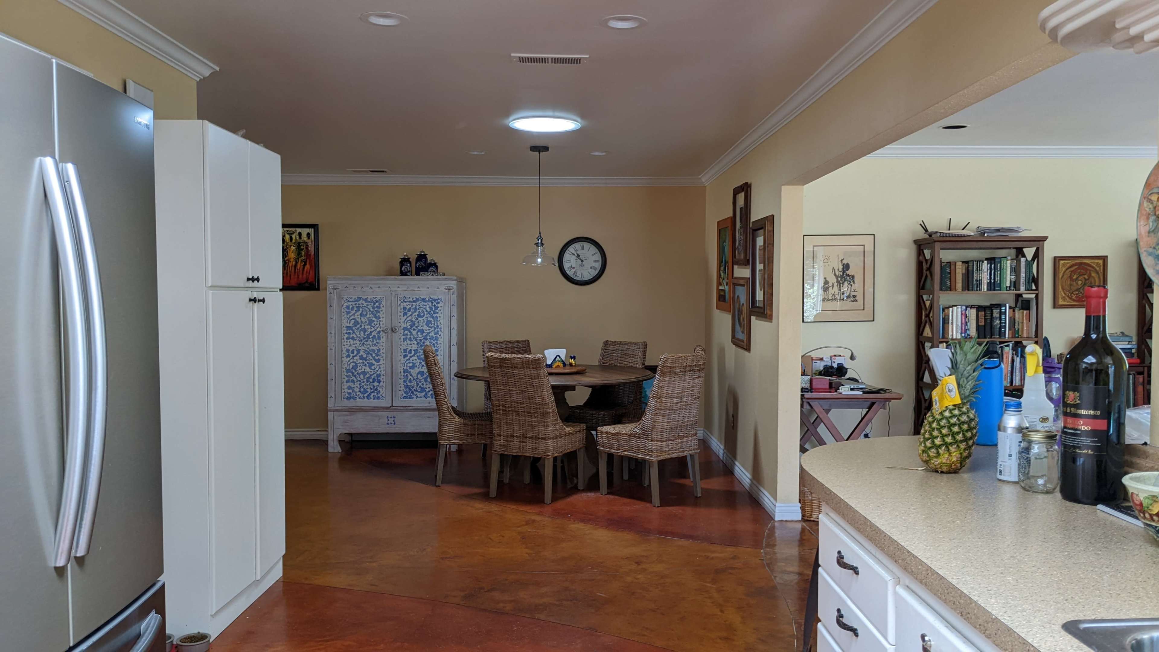 A kitchen opens to a dining area furnished with a round table and four wicker chairs, featuring artwork on the walls and various household items on the countertops.