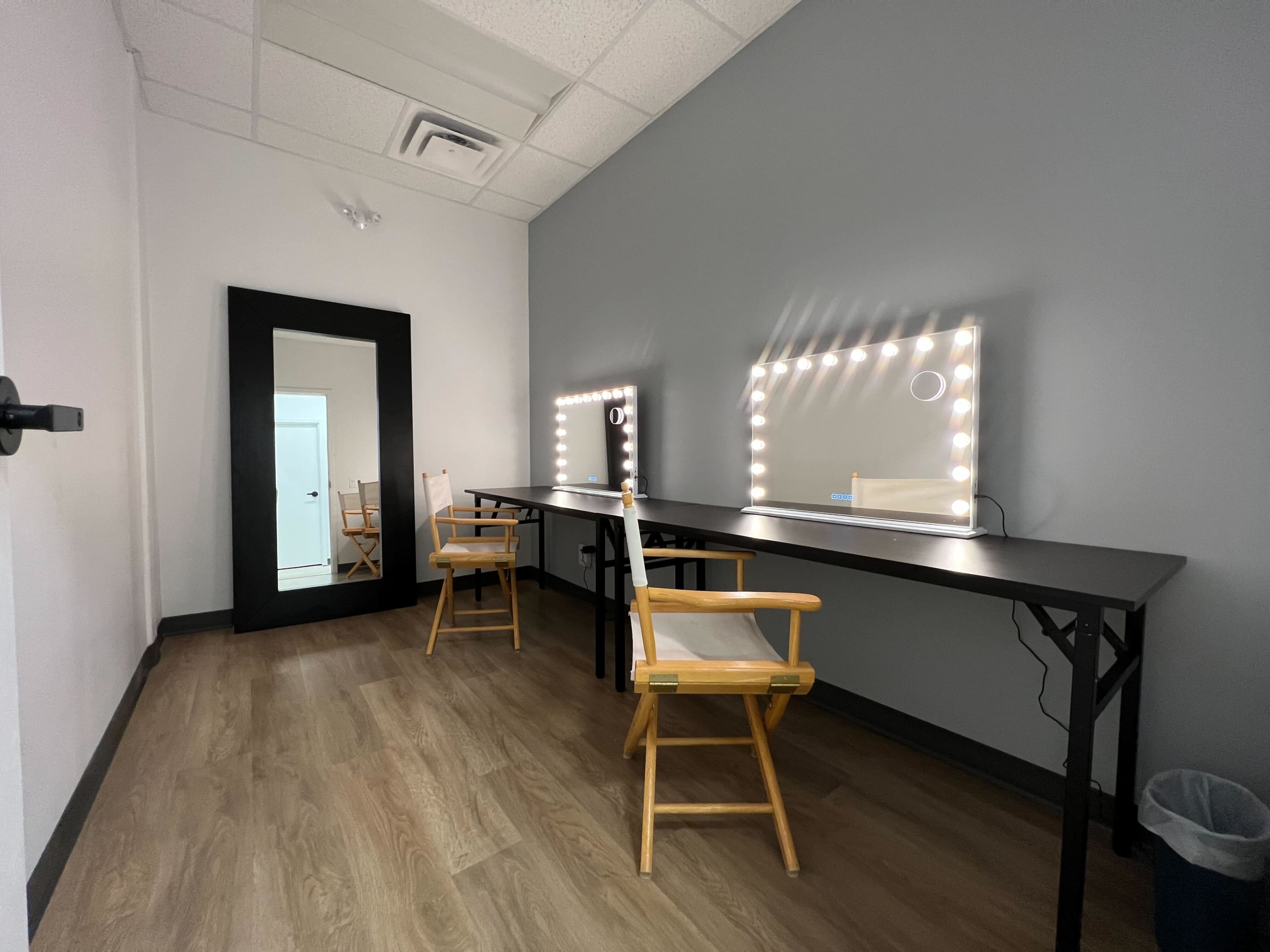 A simple makeup room featuring two lighted mirrors, two chairs, and a full-length mirror against a gray wall.