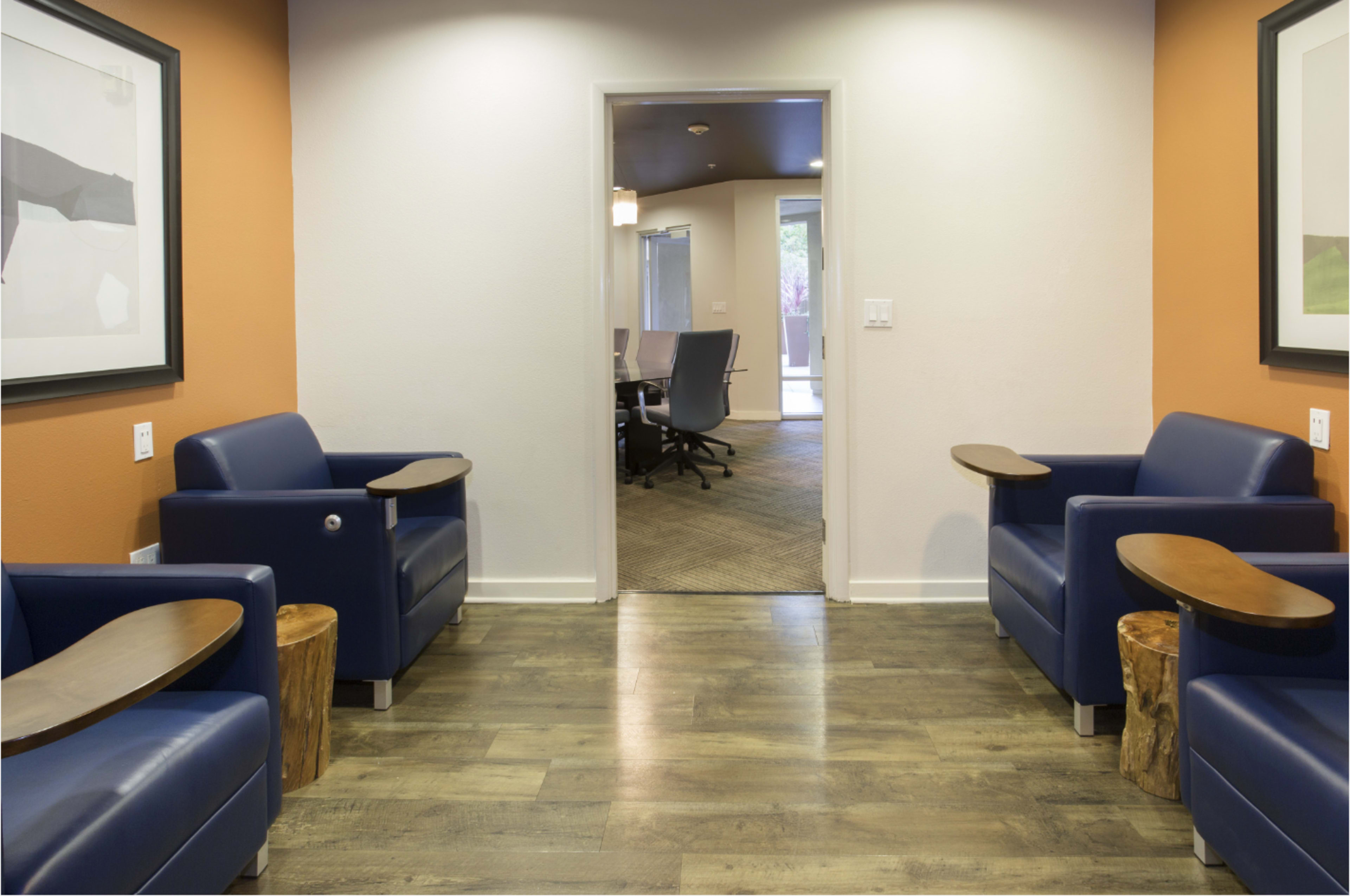 The image shows a modern waiting area with two blue chairs featuring wooden armrests, positioned on a wooden floor, leading to an open doorway with a conference room visible in the background.