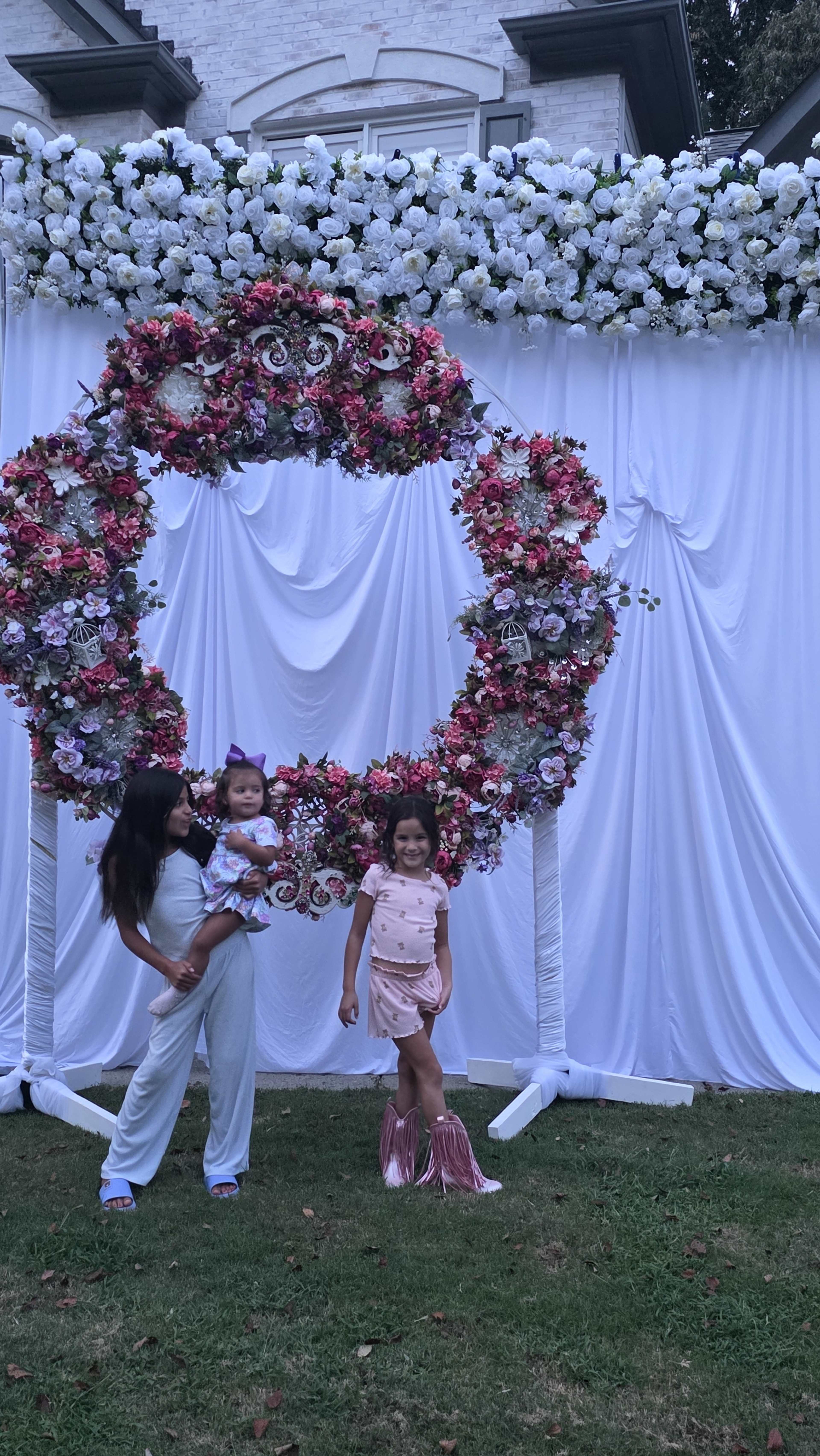 Two girls pose in front of a floral wreath backdrop at an outdoor event.