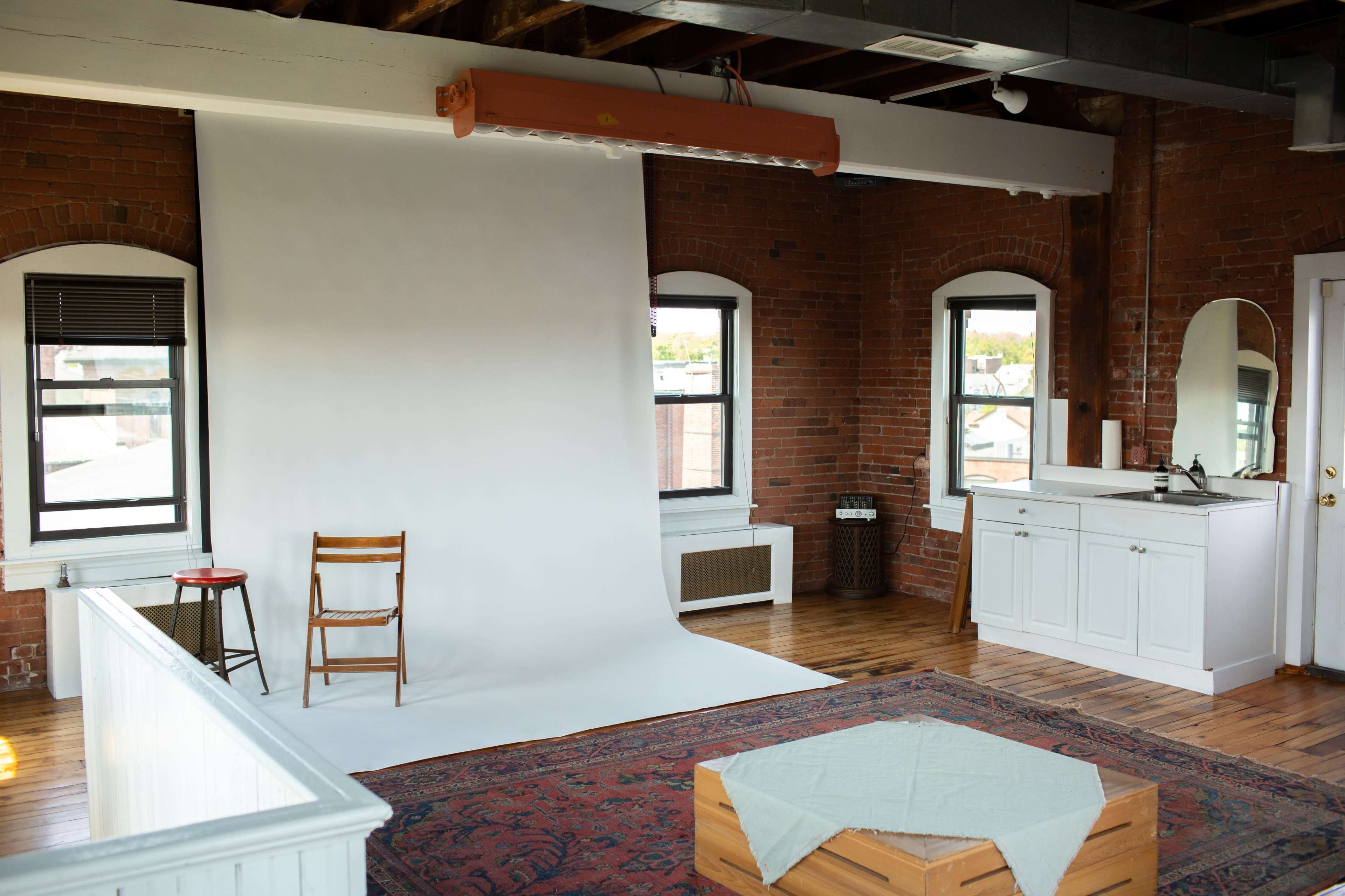 A bright studio space with a white backdrop, a single wooden chair, large windows, and a wooden floor.