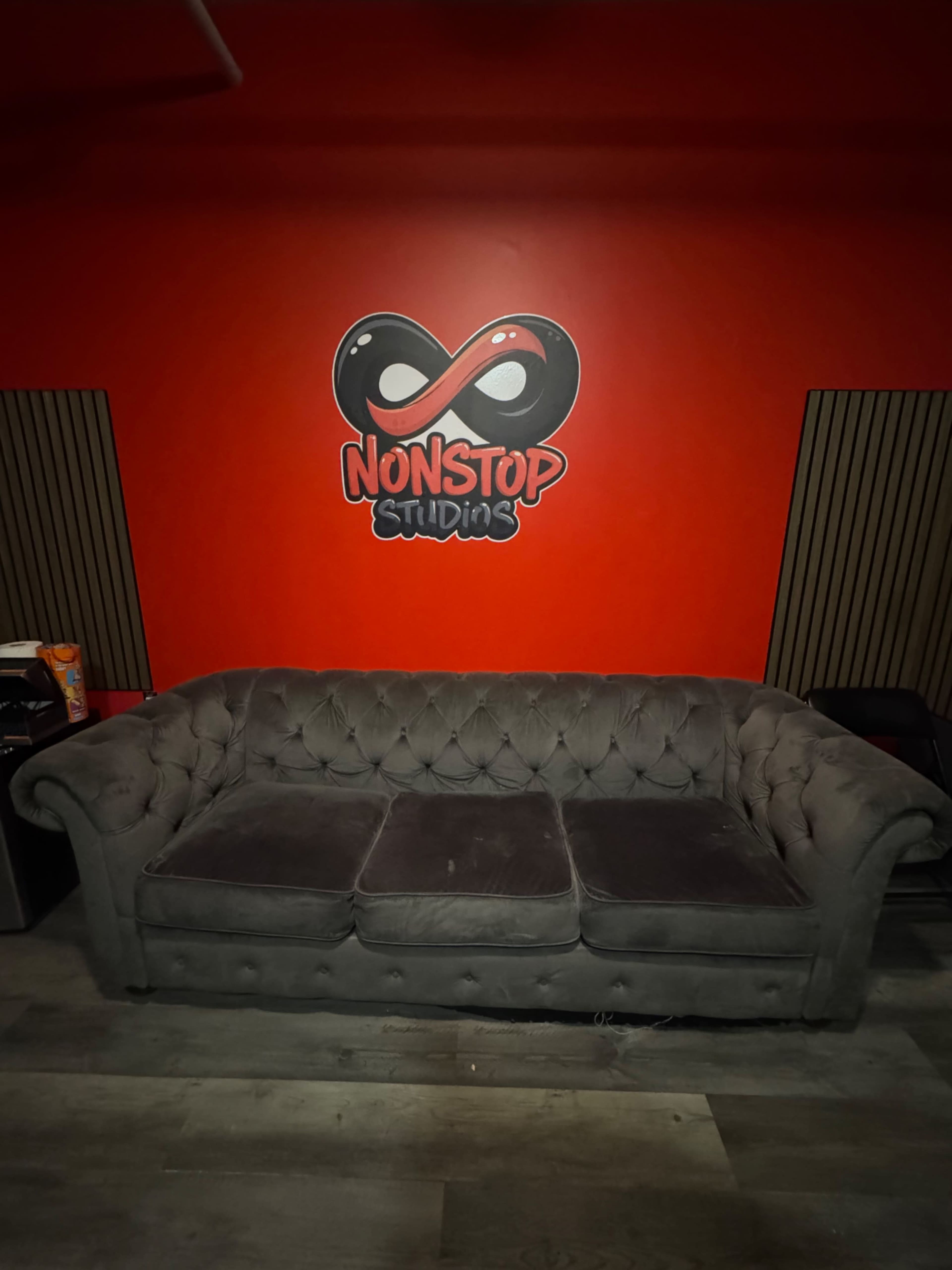 The image shows a gray tufted couch in front of a red wall featuring the logo for "NONSTOP Studios."