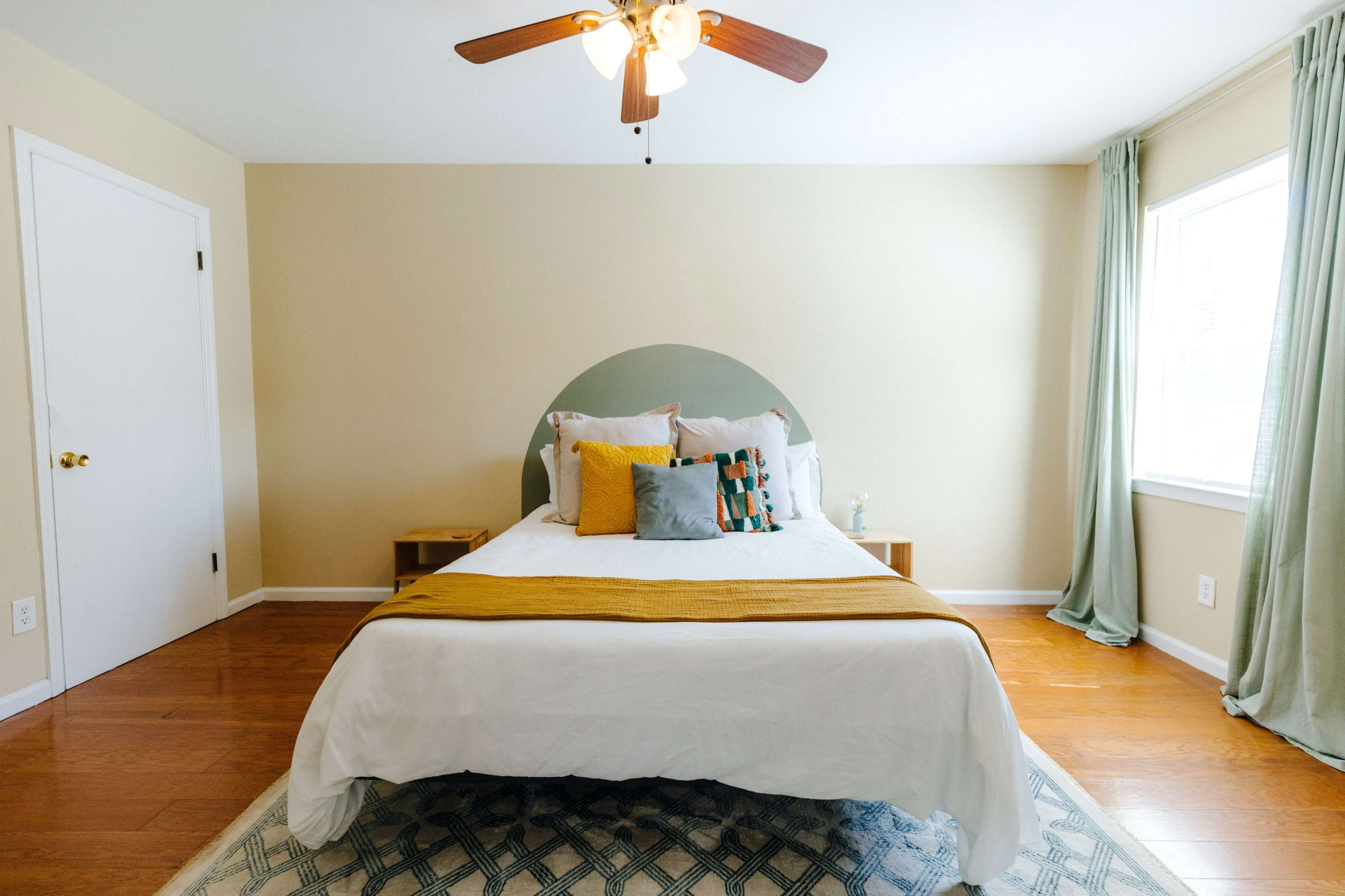 A bedroom features a bed with decorative pillows, a wooden floor, and a ceiling fan, accompanied by a window with curtains.