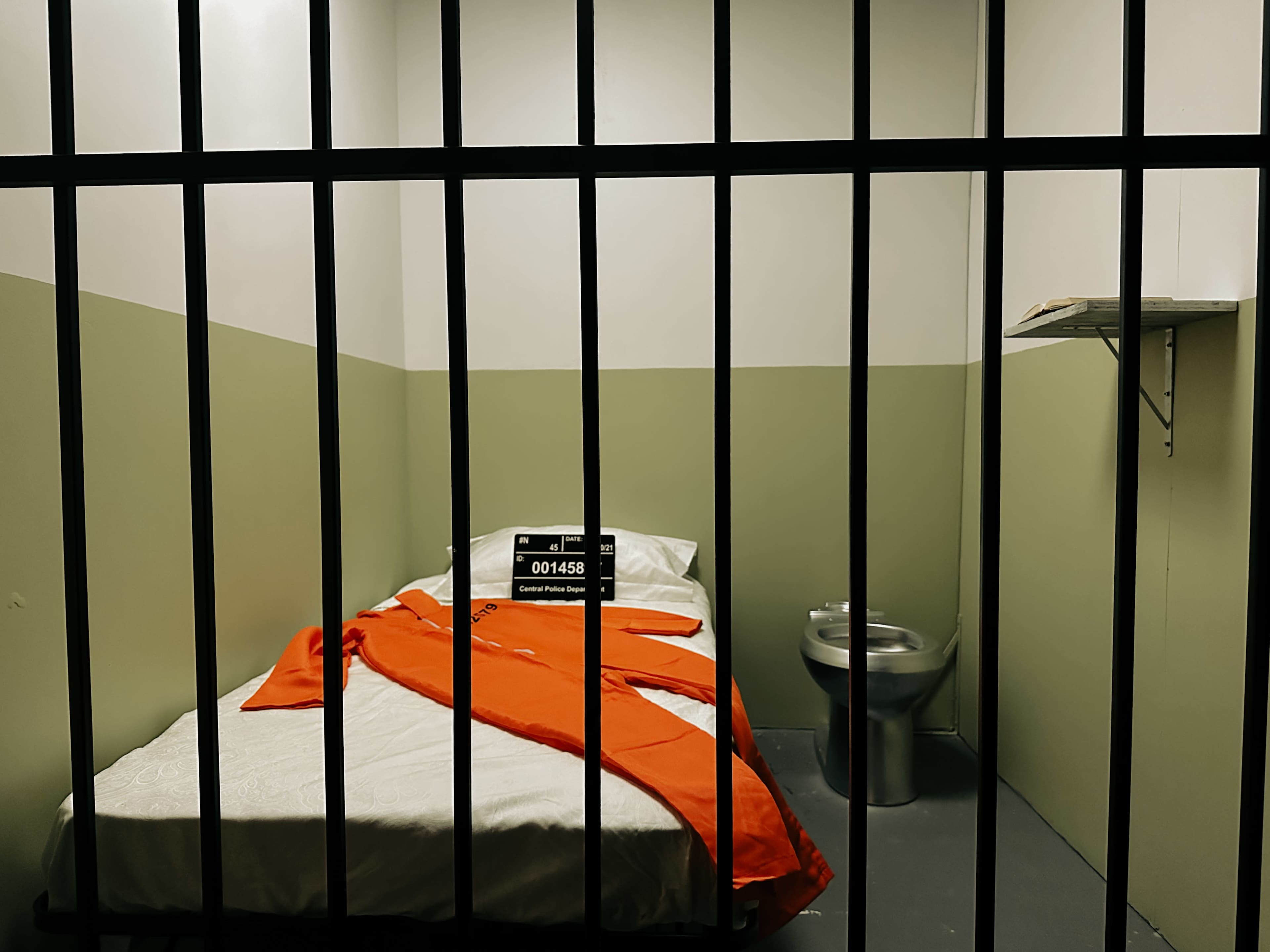 A small, sparse prison cell contains a bed with white linens, a red blanket, a clock, and a toilet.