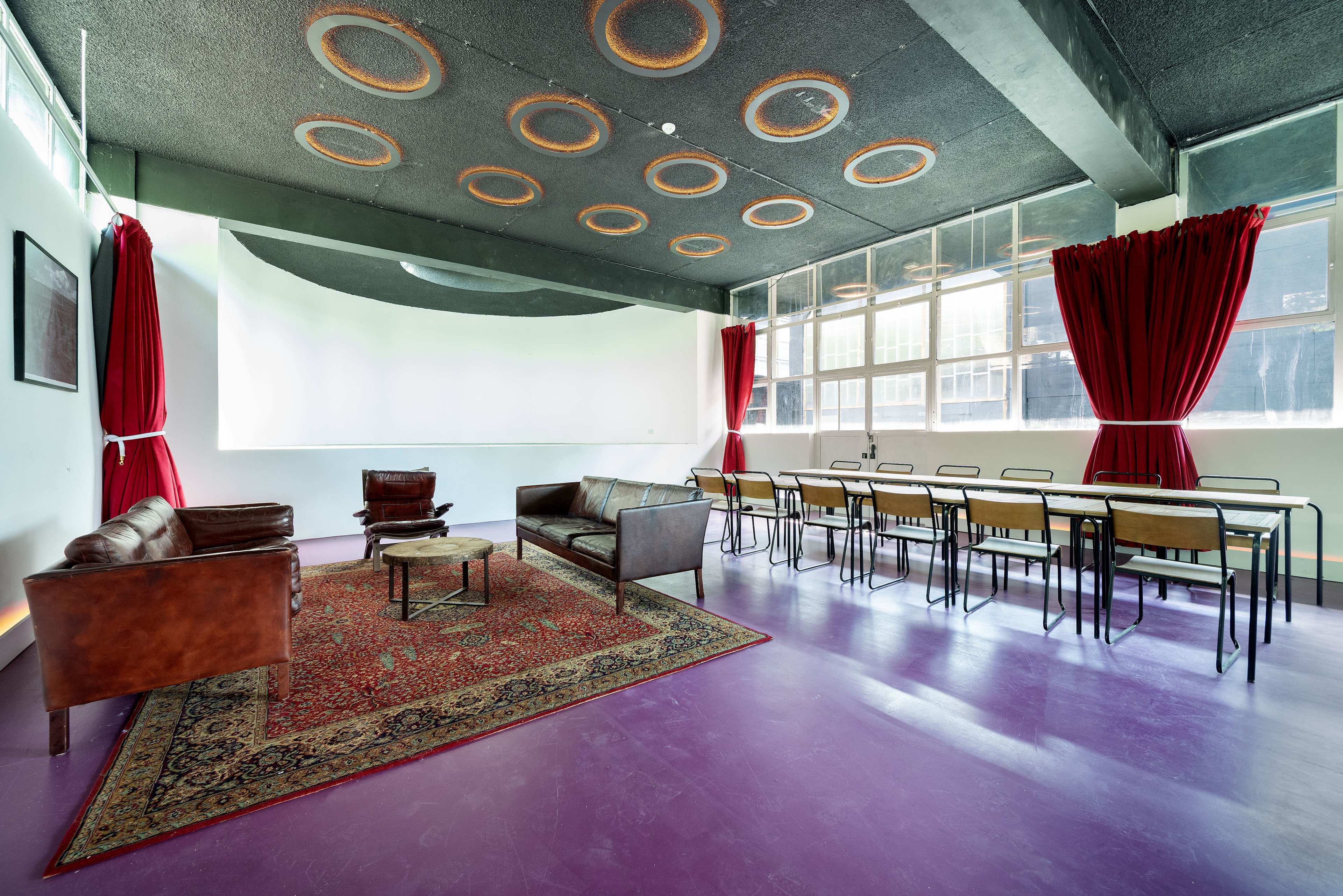 The image features a modern, spacious room with a seating area of leather sofas and chairs, a long table with chairs, large windows, and red curtains.