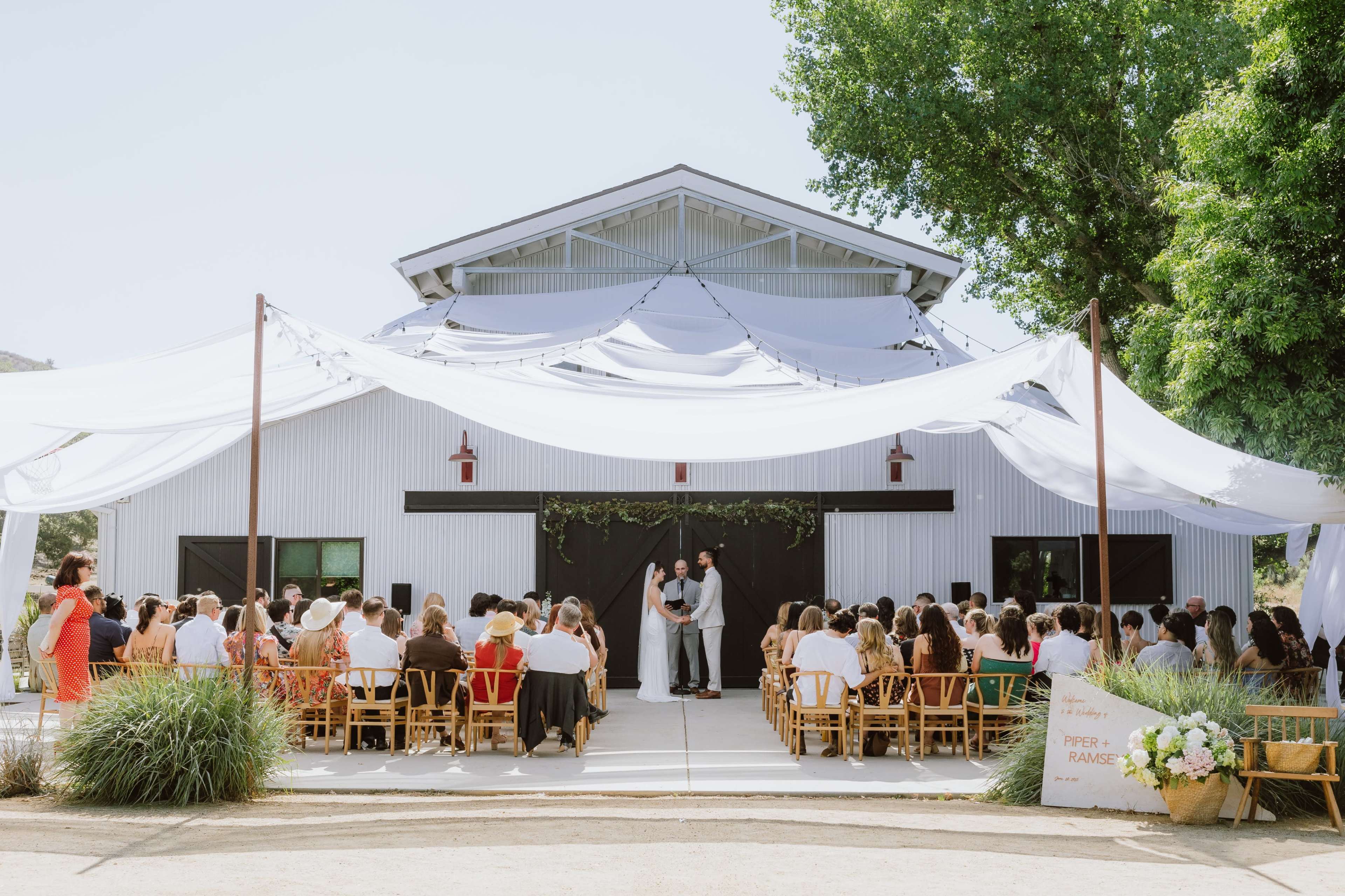 A couple stands under a draped canopy at an outdoor wedding ceremony in front of a barn, surrounded by seated guests.