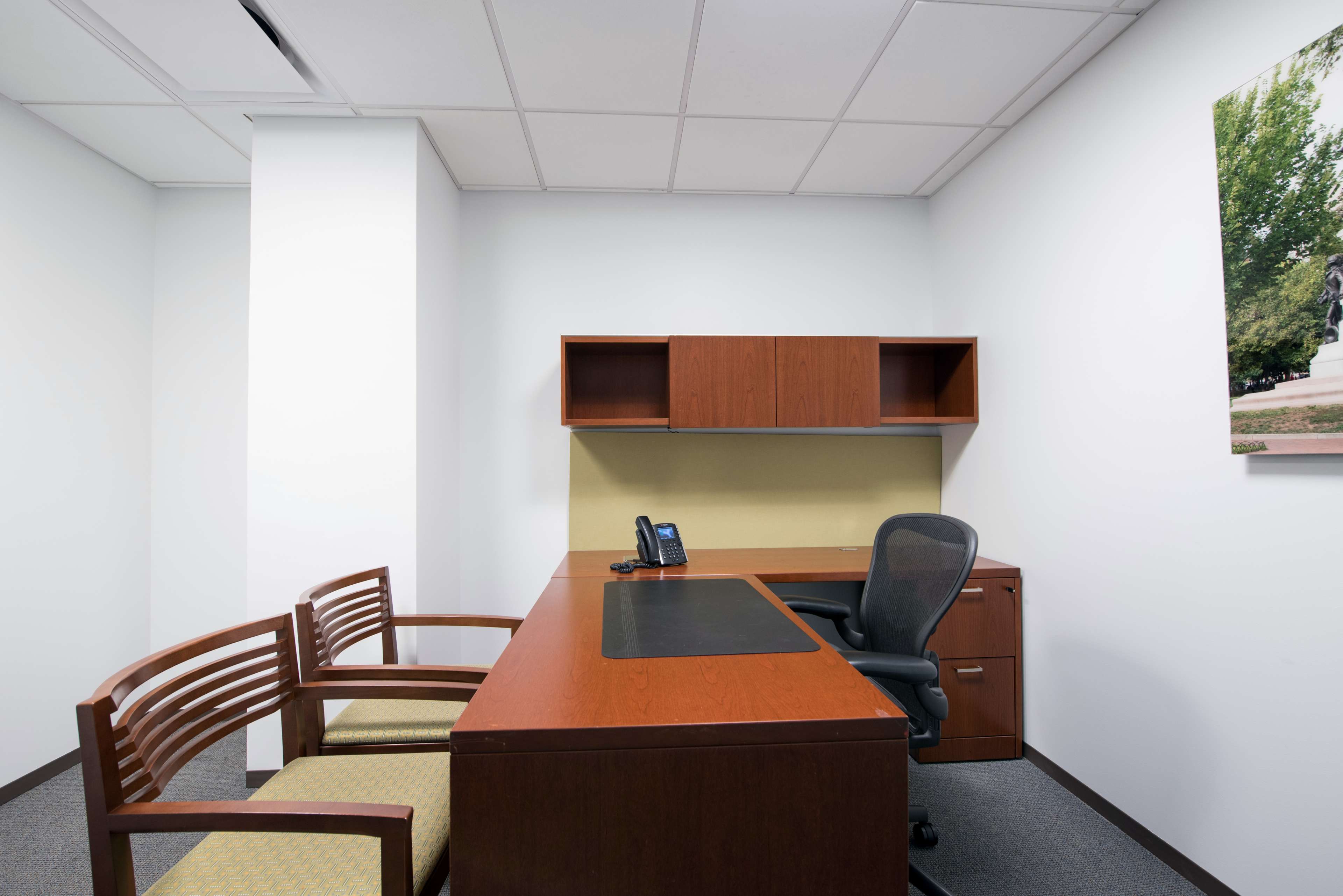 A minimalistic office space featuring a wooden desk, an office chair, and two wooden chairs against a plain wall.