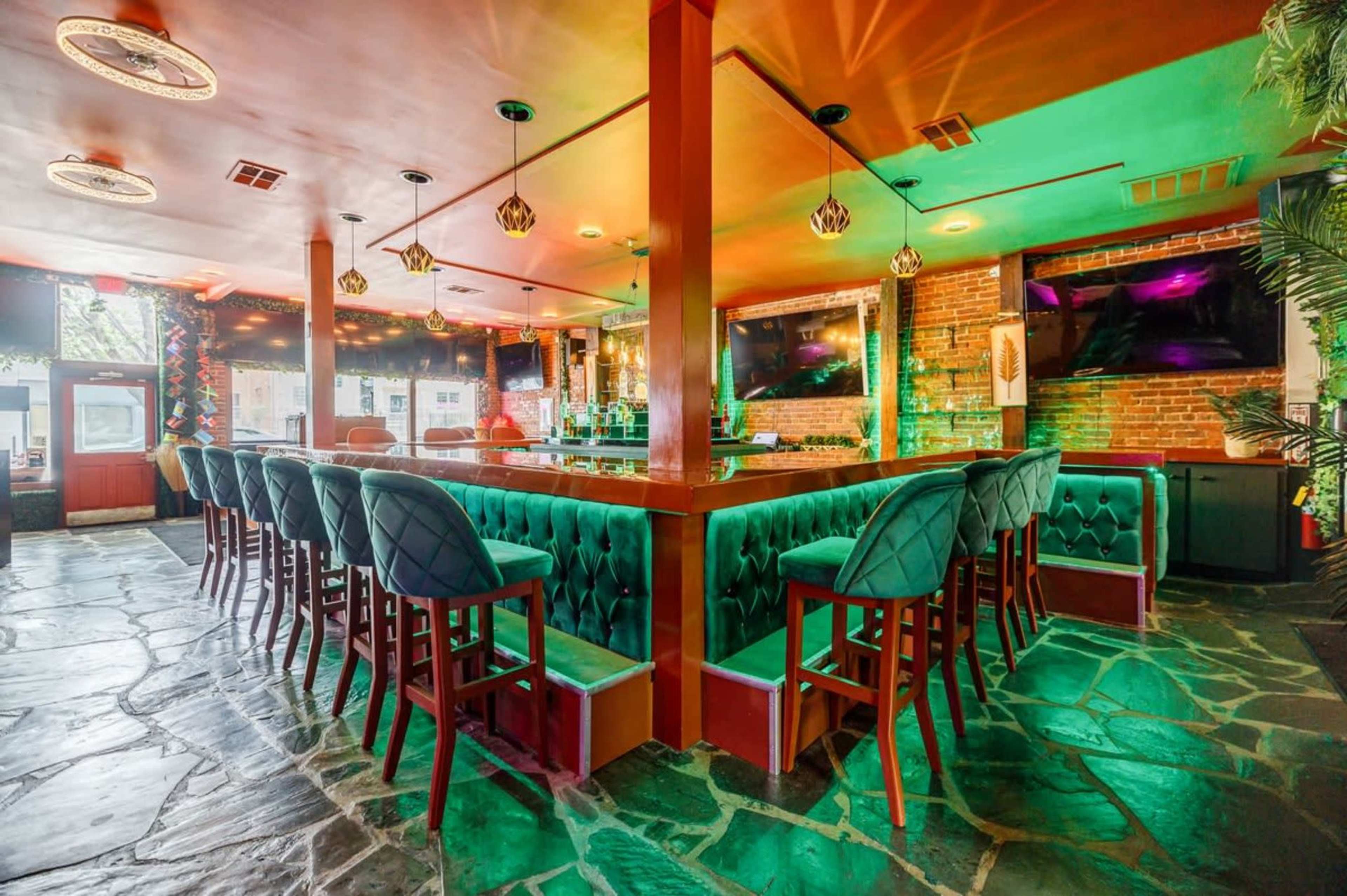 The image shows a modern bar with a green-lit counter, surrounded by velvet-upholstered stools, set against a backdrop of exposed brick walls and colorful ambient lighting.