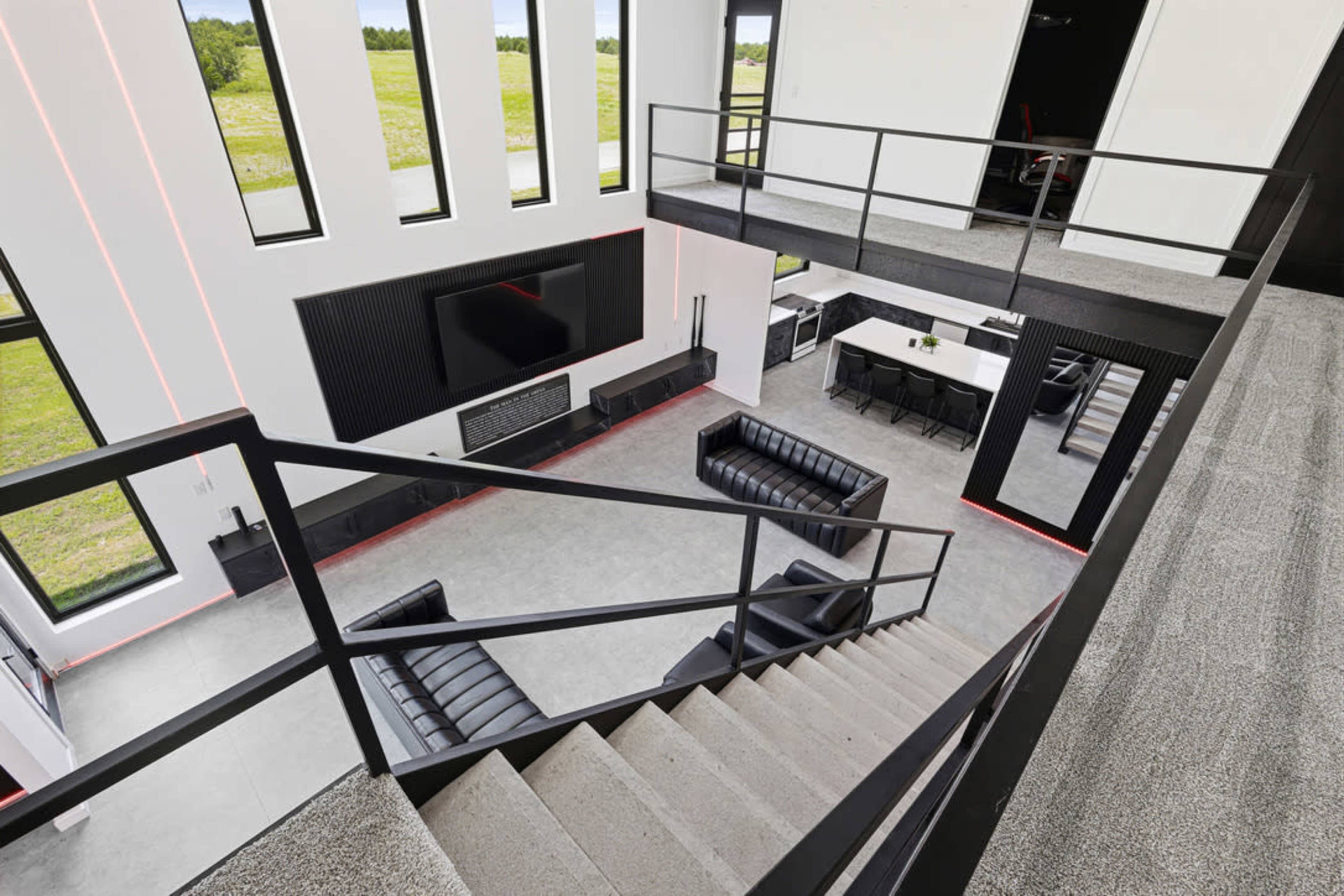 The image showcases a modern, open-concept living area with large windows, black leather furniture, and a staircase leading to a second level.