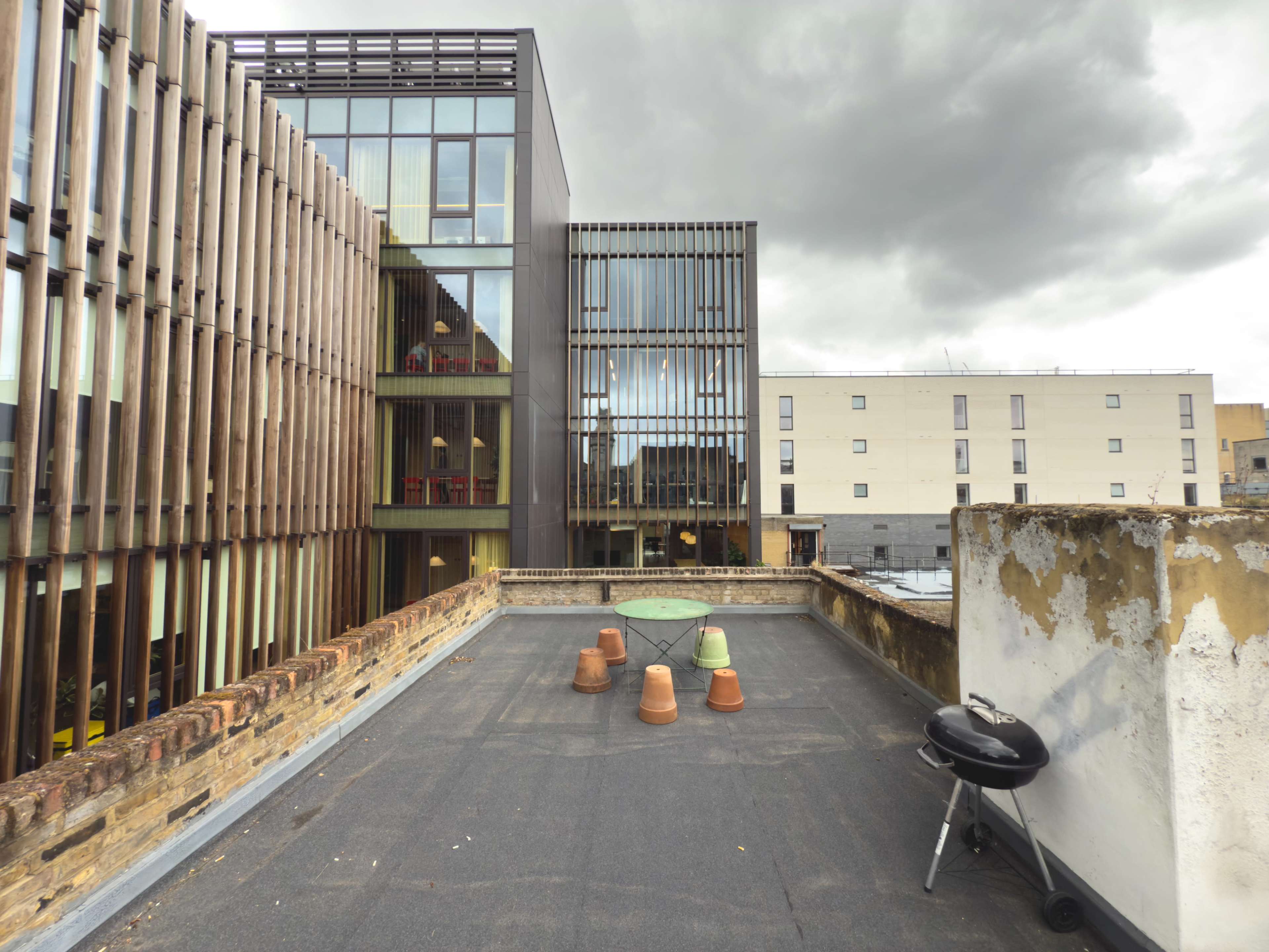 The image shows a rooftop area with terracotta plant pots and a charcoal grill, set against a backdrop of modern buildings and a cloudy sky.