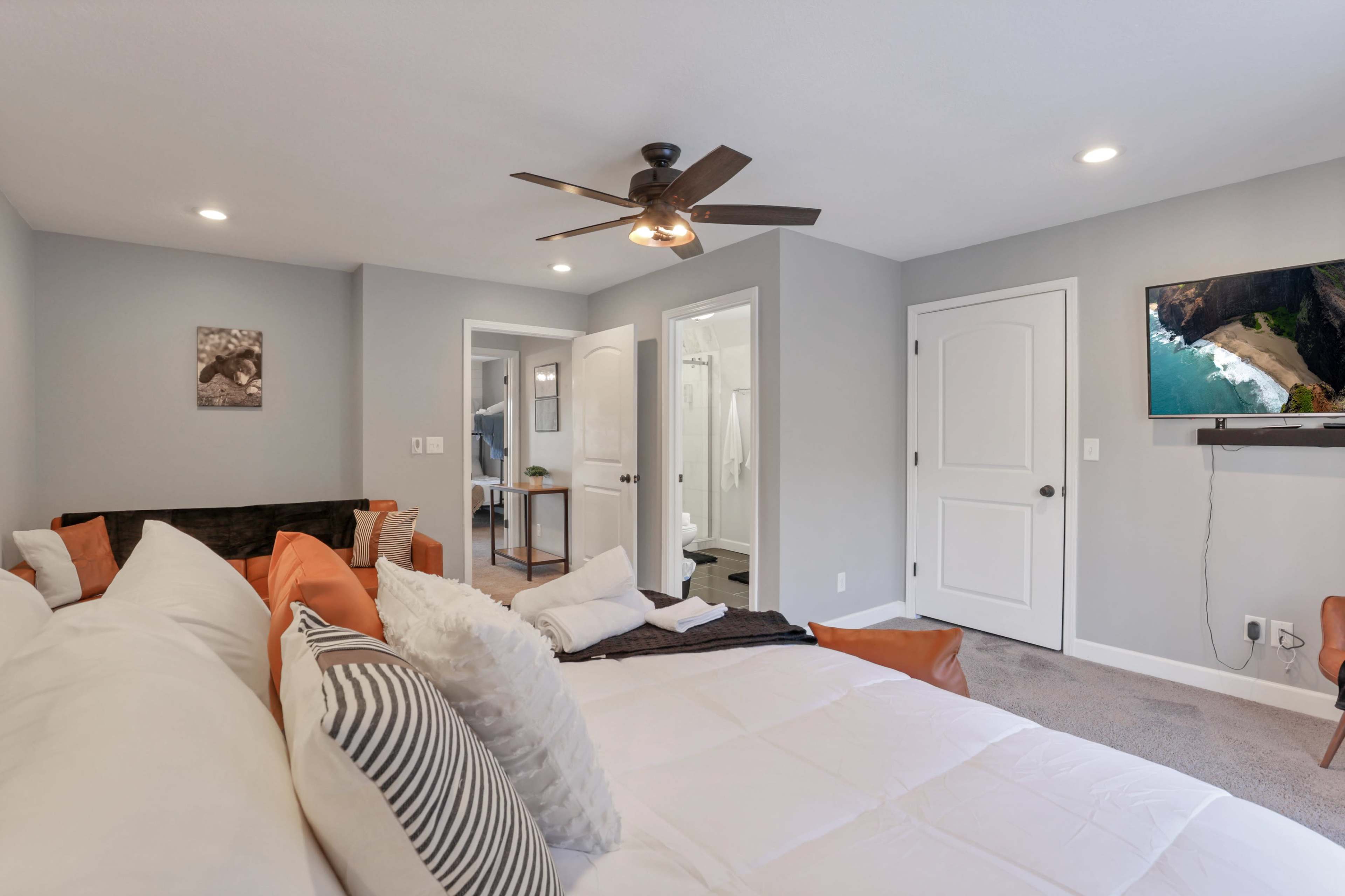 A modern bedroom featuring a large bed with white bedding, a ceiling fan, and an attached bathroom visible in the background.