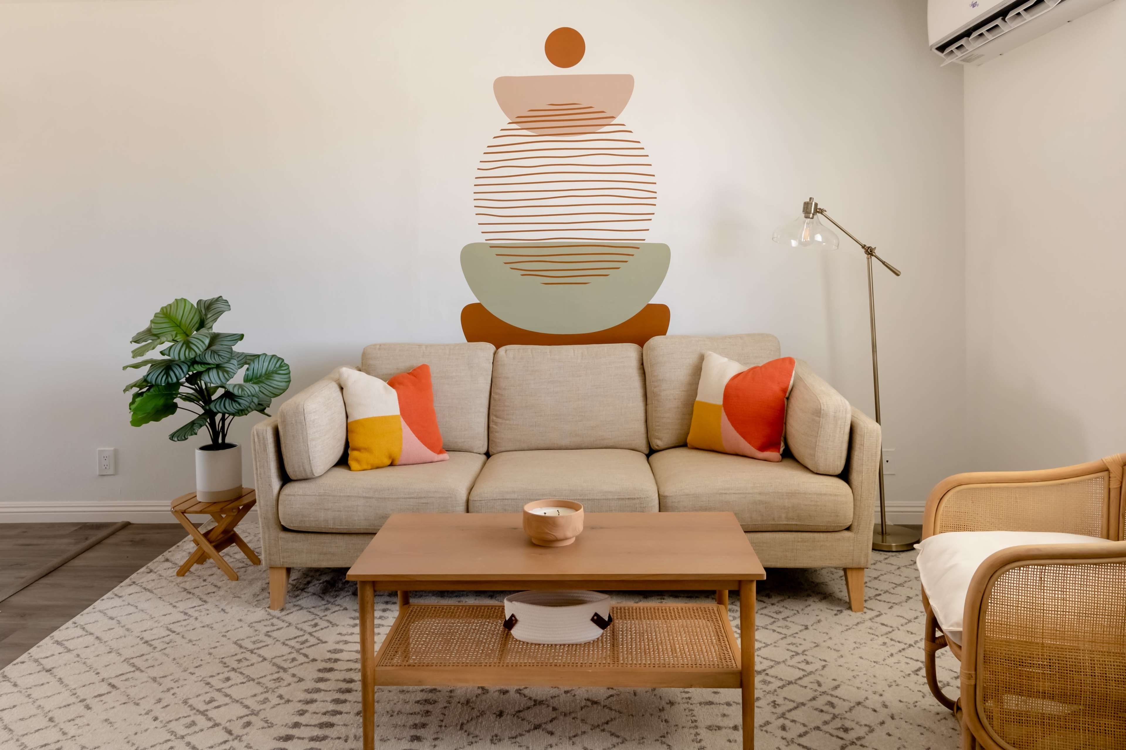 A light-colored sofa with colorful pillows is positioned in a room featuring a decorative wall mural and a wooden coffee table.