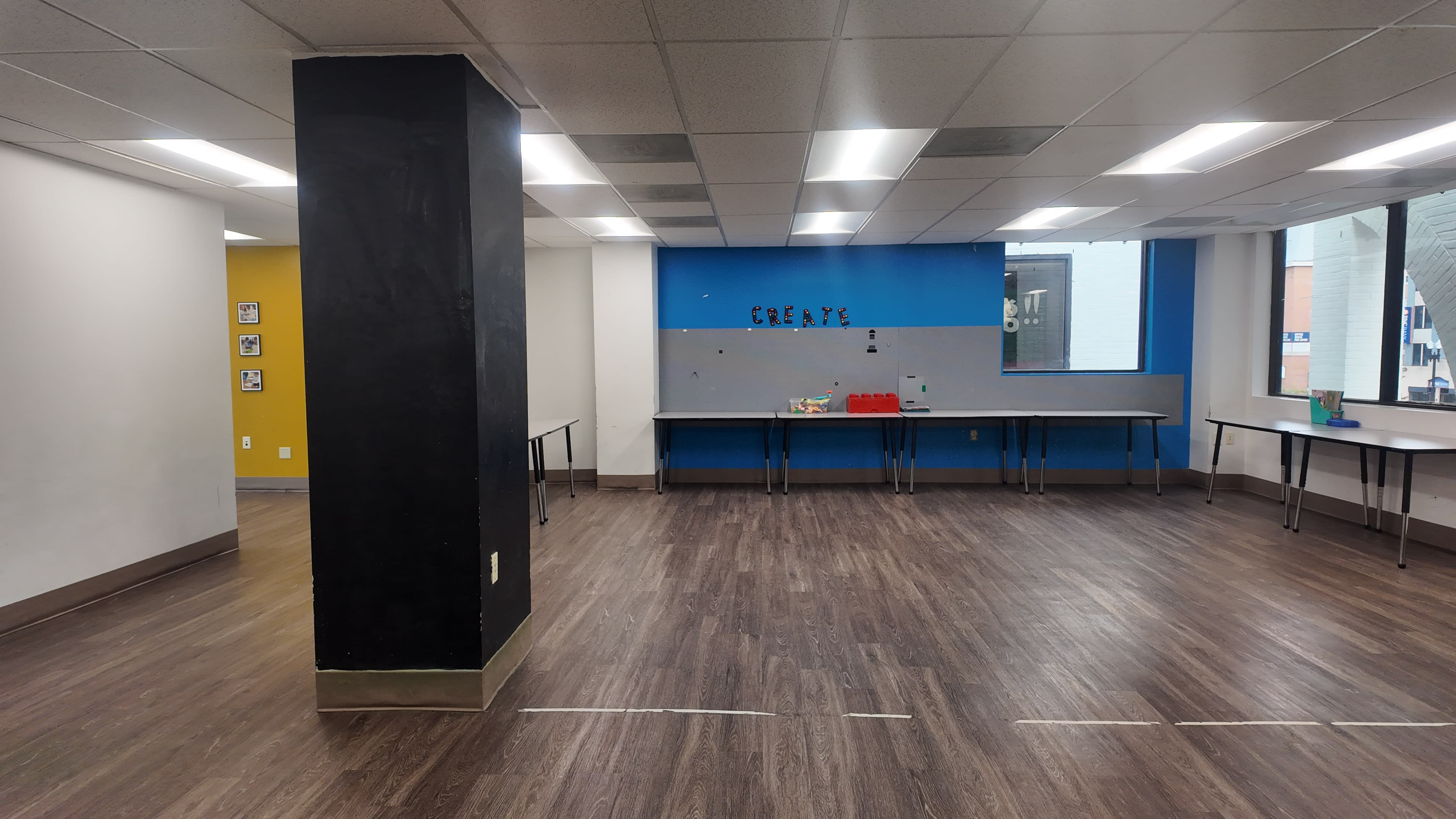 The image shows an empty room with wooden flooring, a chalkboard column, and tables along the walls, featuring blue and yellow accent colors.