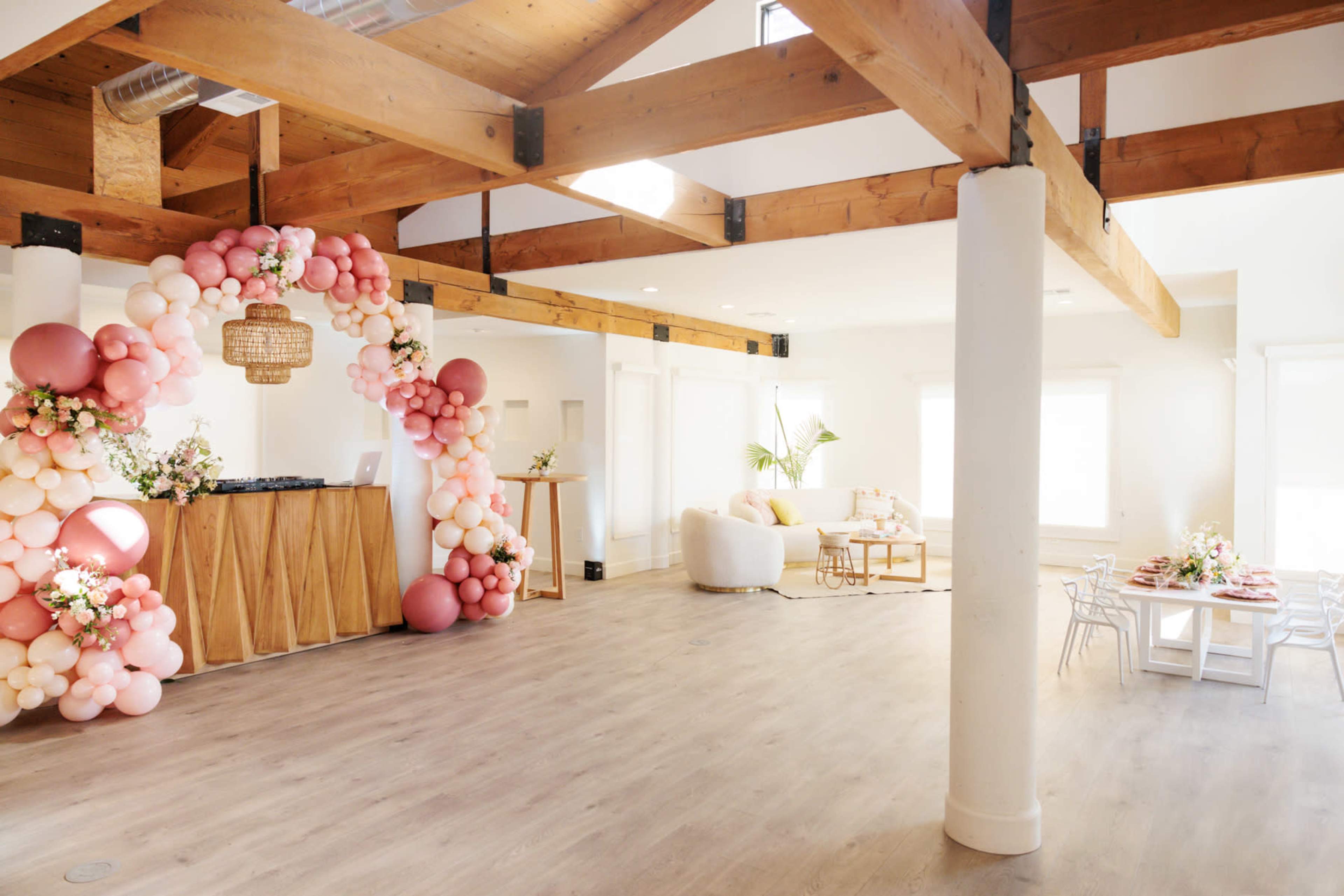 A bright interior space with wooden beams, featuring a decorative balloon arch to one side, a bar area, and a seating arrangement with a large white sofa and dining table.