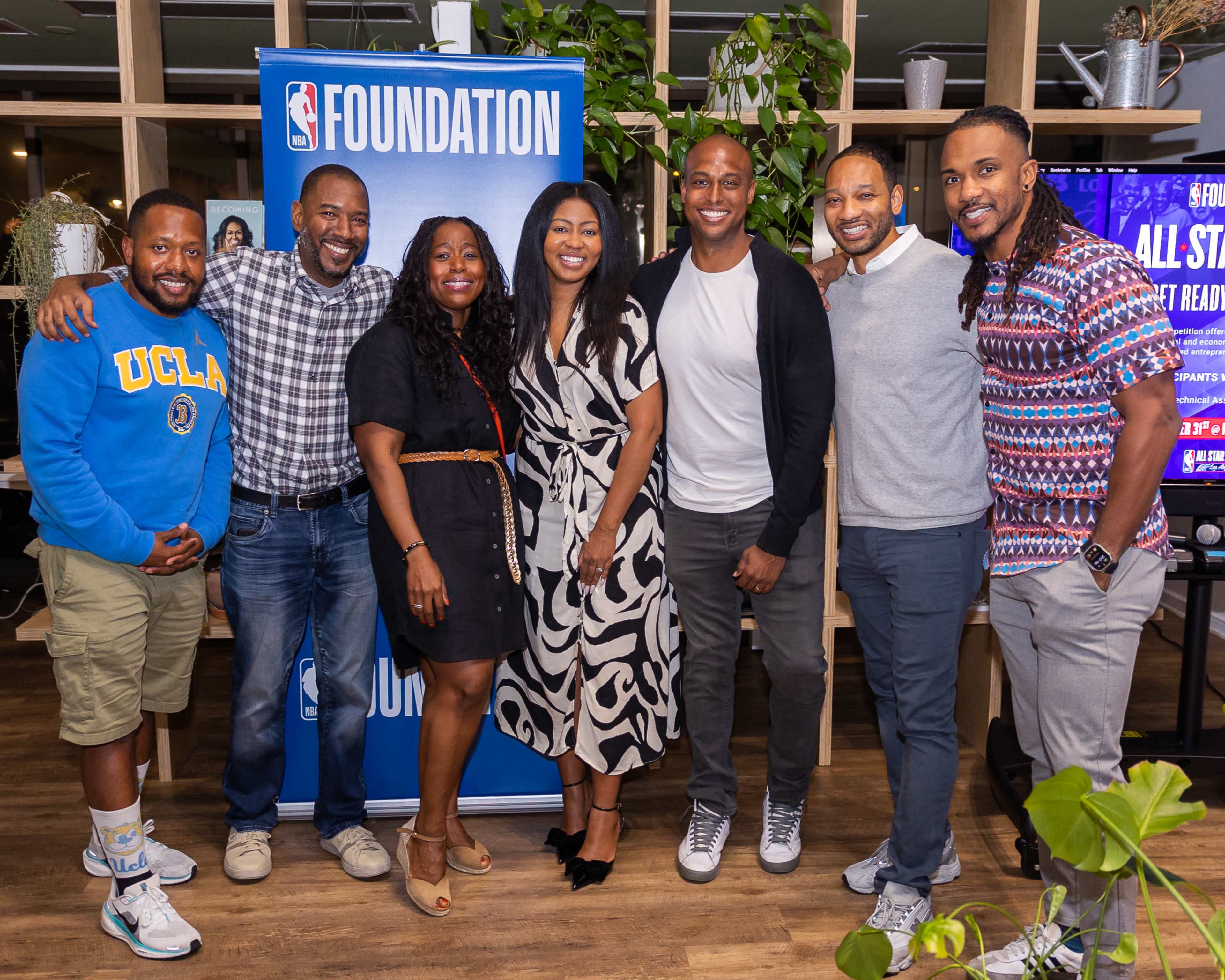 A group of seven people poses for a photo in front of an NBA Foundation banner, inside a well-decorated indoor space with plants.