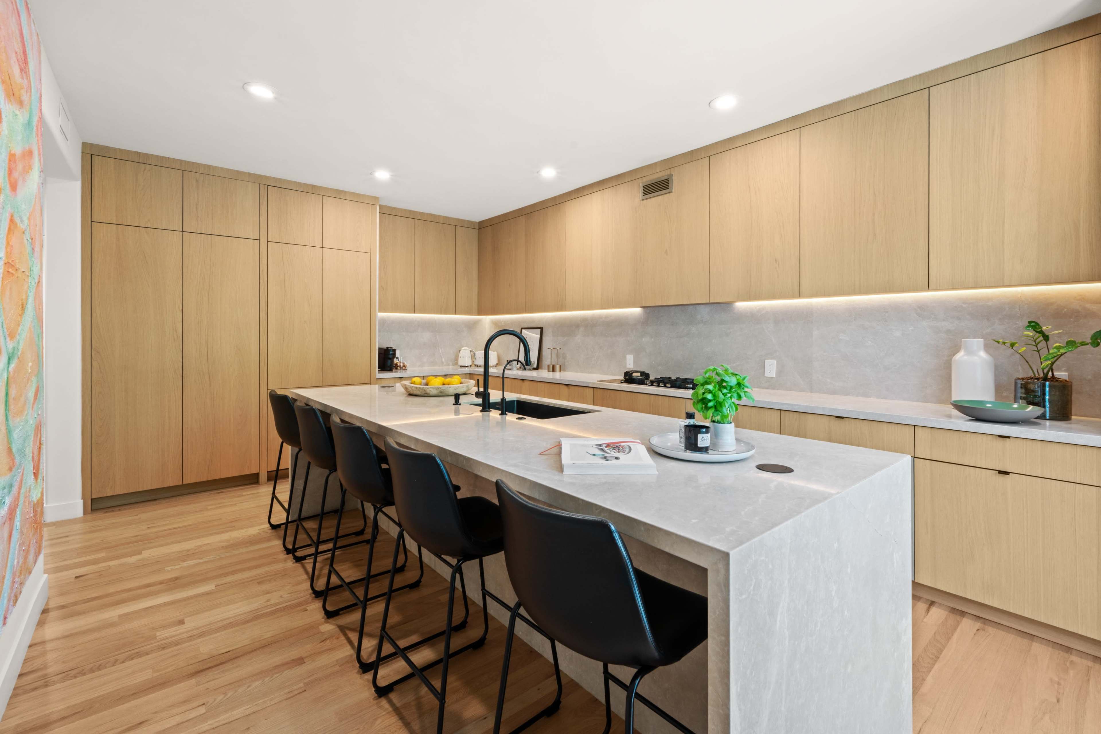 The image shows a modern kitchen with light wood cabinetry, a large stone island with seating, and recessed lighting.
