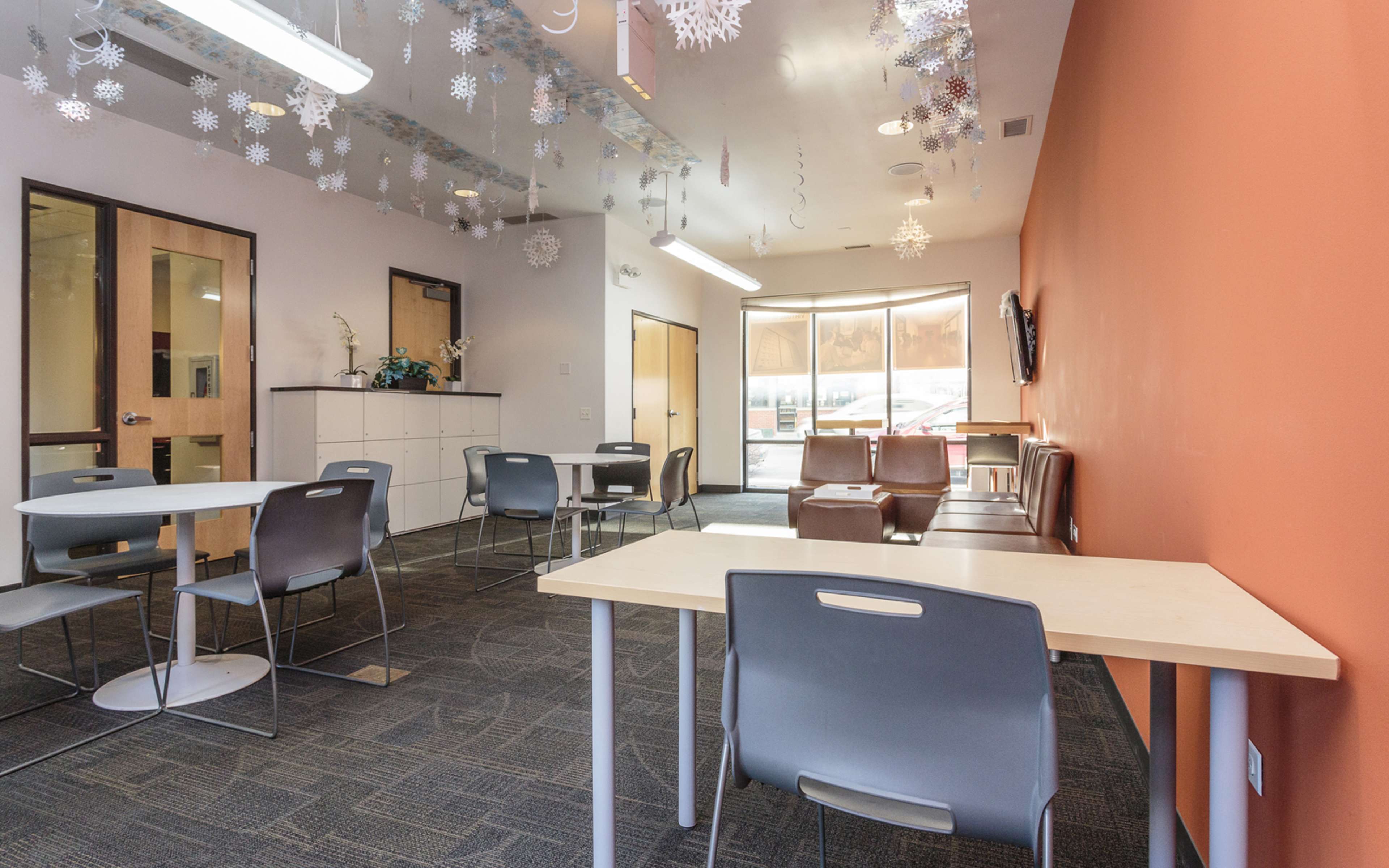 The room features several tables and chairs arranged on a carpeted floor under hanging snowflake decorations, with large windows allowing natural light.