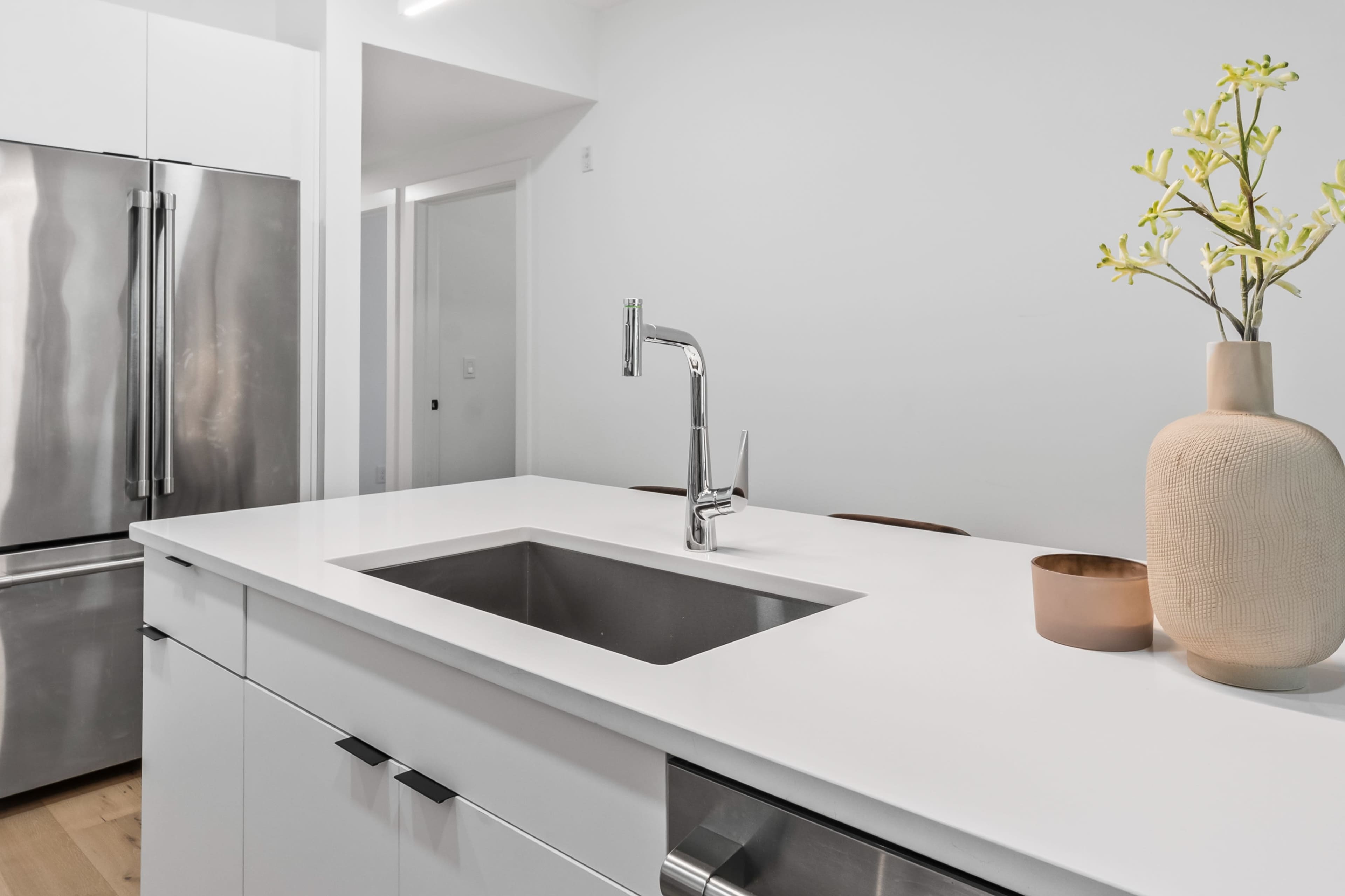 The image shows a modern kitchen with a stainless steel sink and faucet on a white countertop, accompanied by a beige vase and a small bowl.