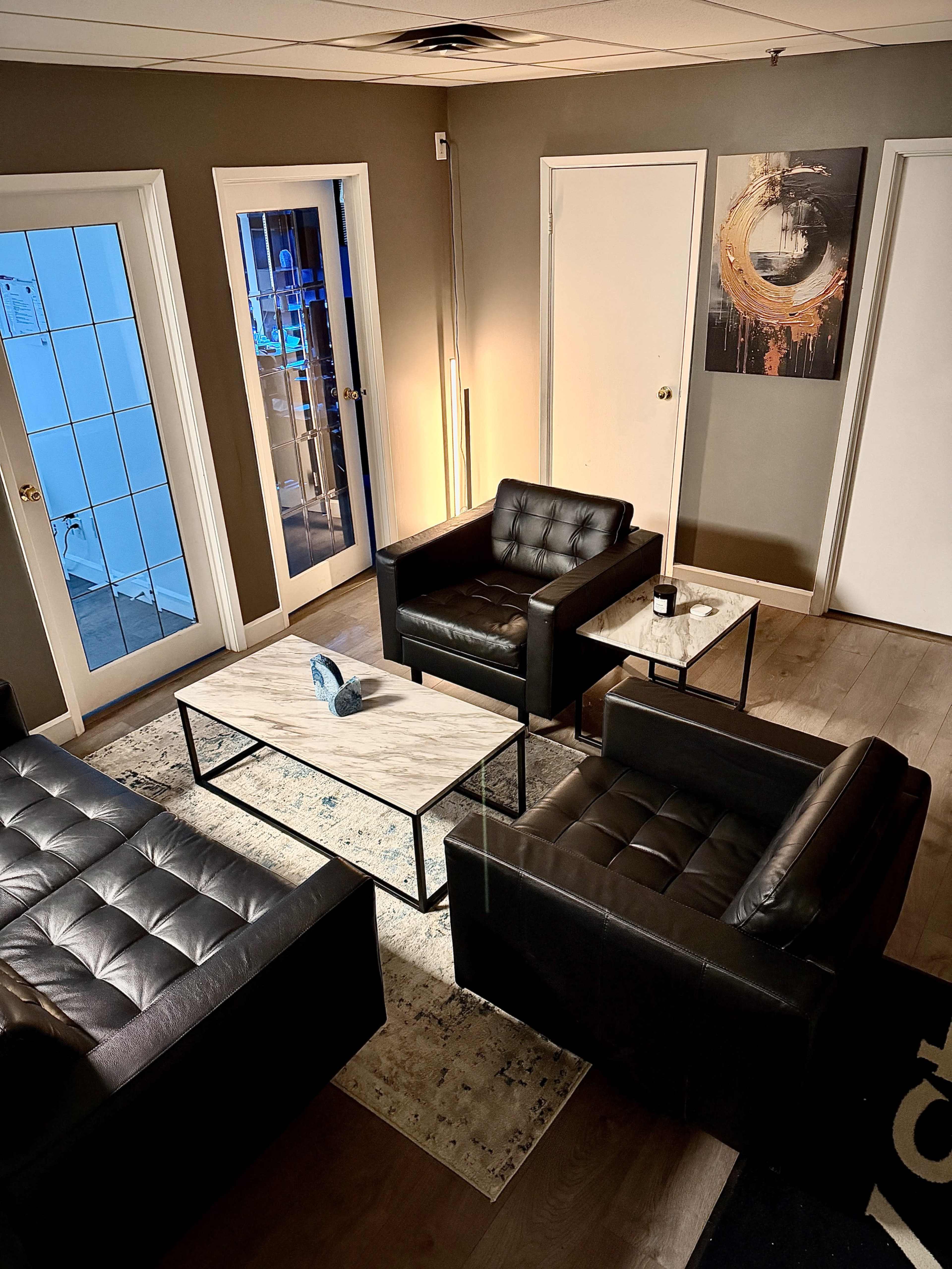 The image shows a modern lounge area with two black leather chairs, a marble-topped coffee table, and framed artwork on the wall, illuminated by artificial light.