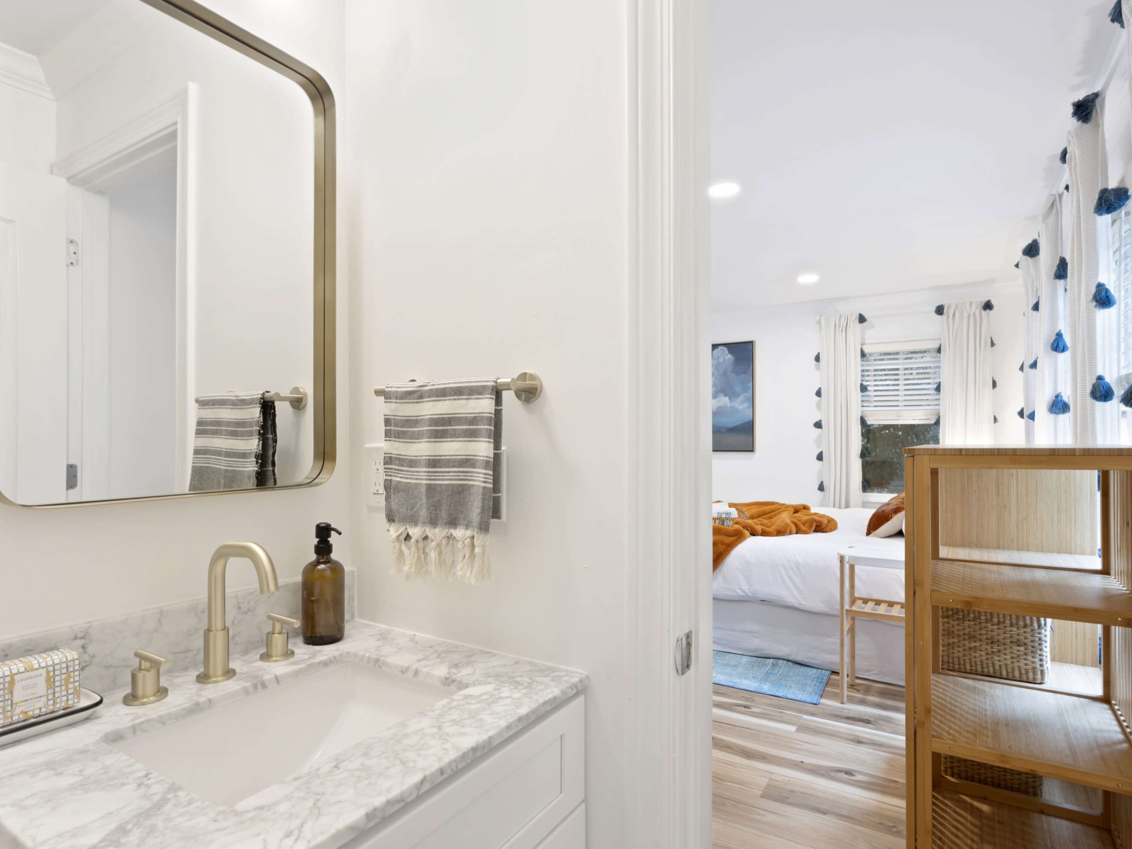 A bathroom featuring a marble countertop sink with a mirror, adjacent to a bedroom with a bed covered in blankets and natural light streaming through the windows.