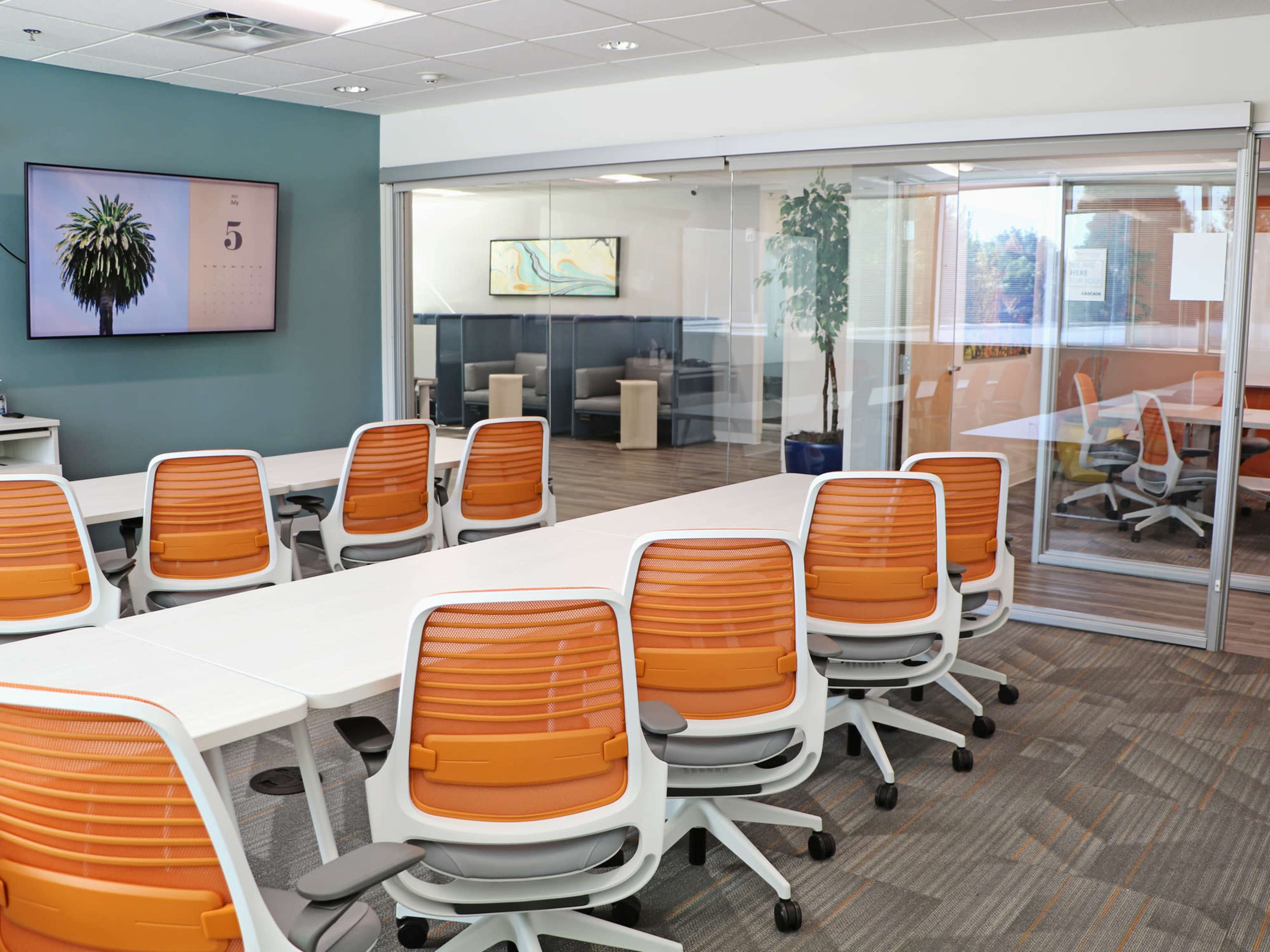 A modern conference room with white tables and orange office chairs, featuring glass walls and a large wall-mounted monitor displaying an image of a plant.
