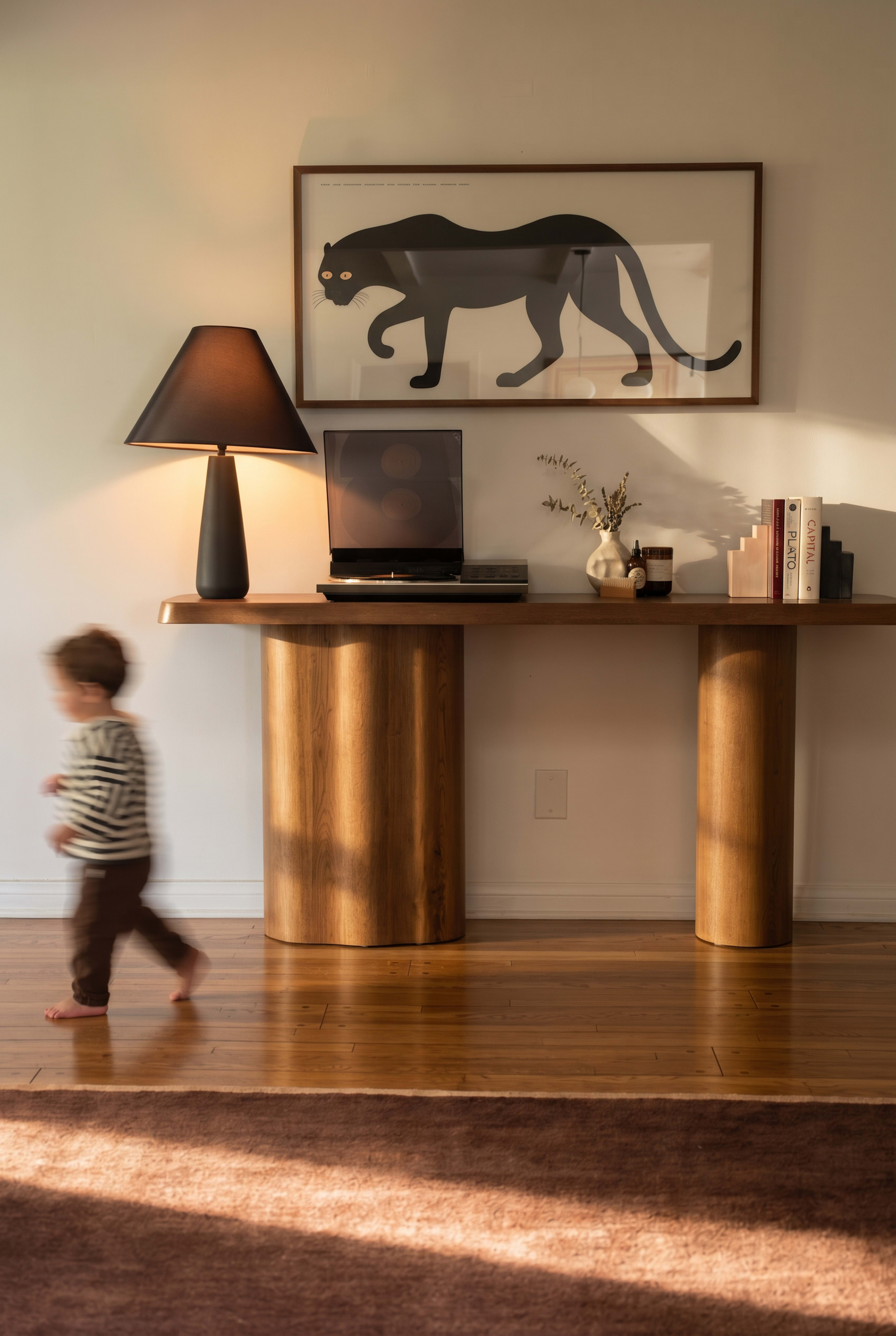 A young child walks past a wooden console table with a laptop, a lamp, and decorative items, while a framed artwork of a panther hangs on the wall.