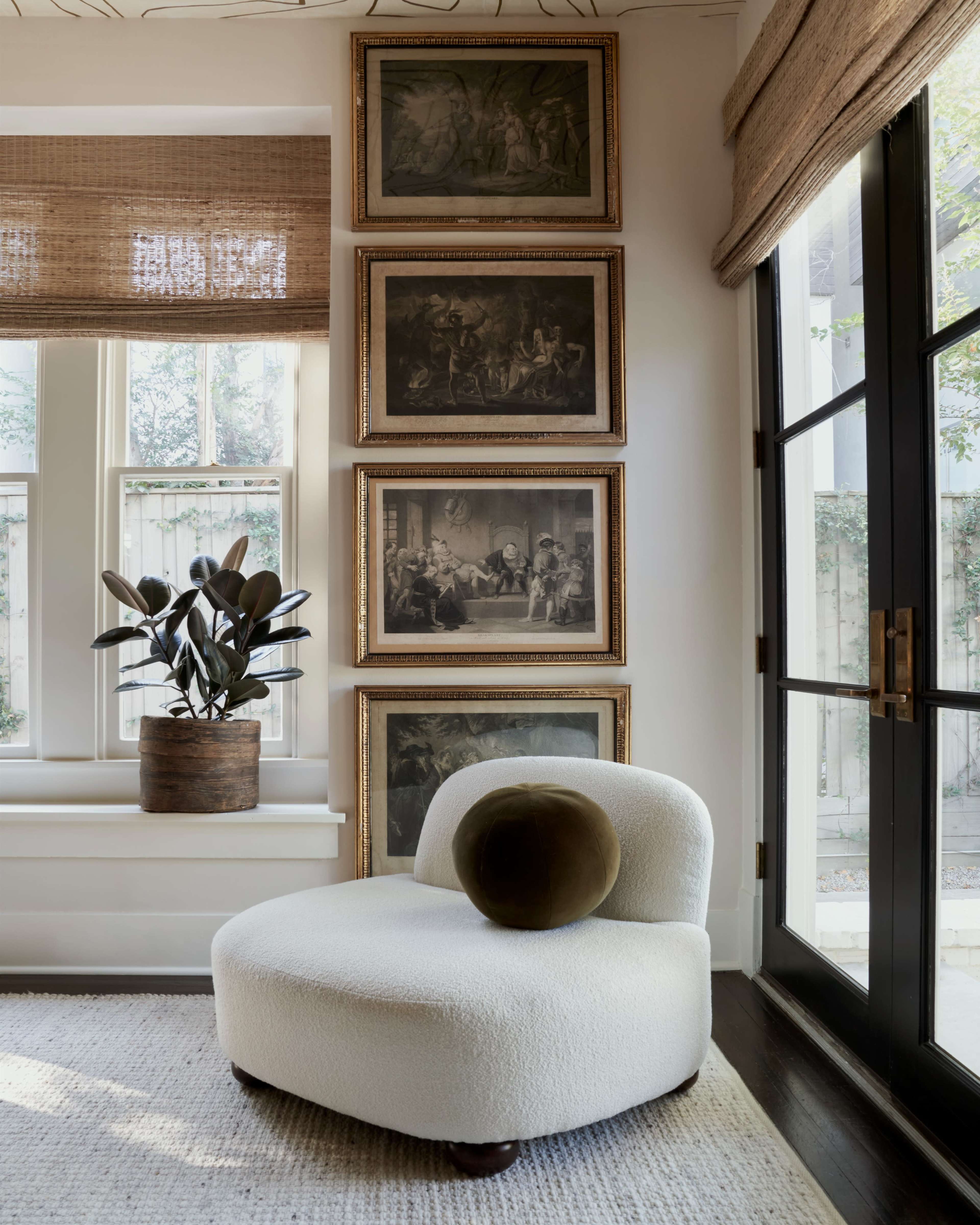 A white upholstered chair with a round green pillow sits next to tall windows adorned with woven shades and framed artwork hung on the wall.