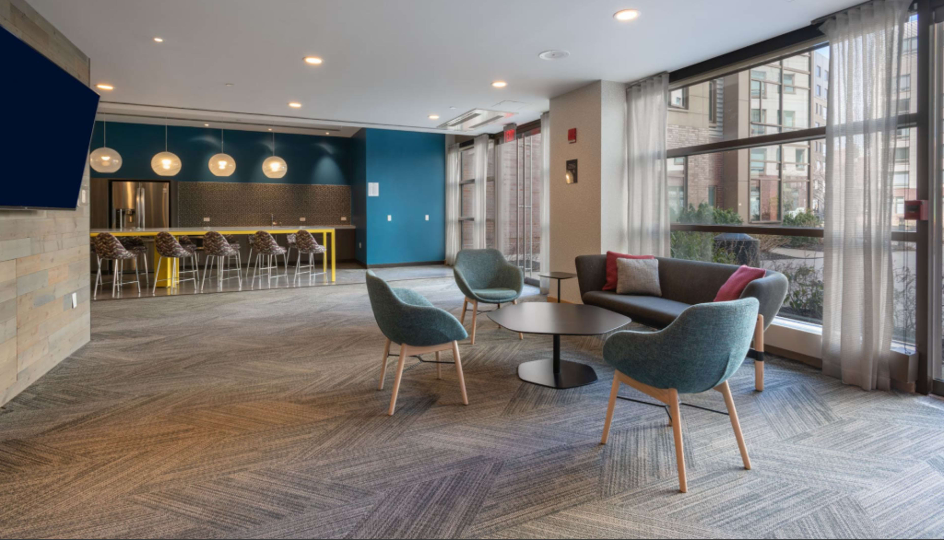The image shows a modern lounge area with a gray couch, two blue chairs, and a round table, alongside a kitchenette and large windows providing natural light.