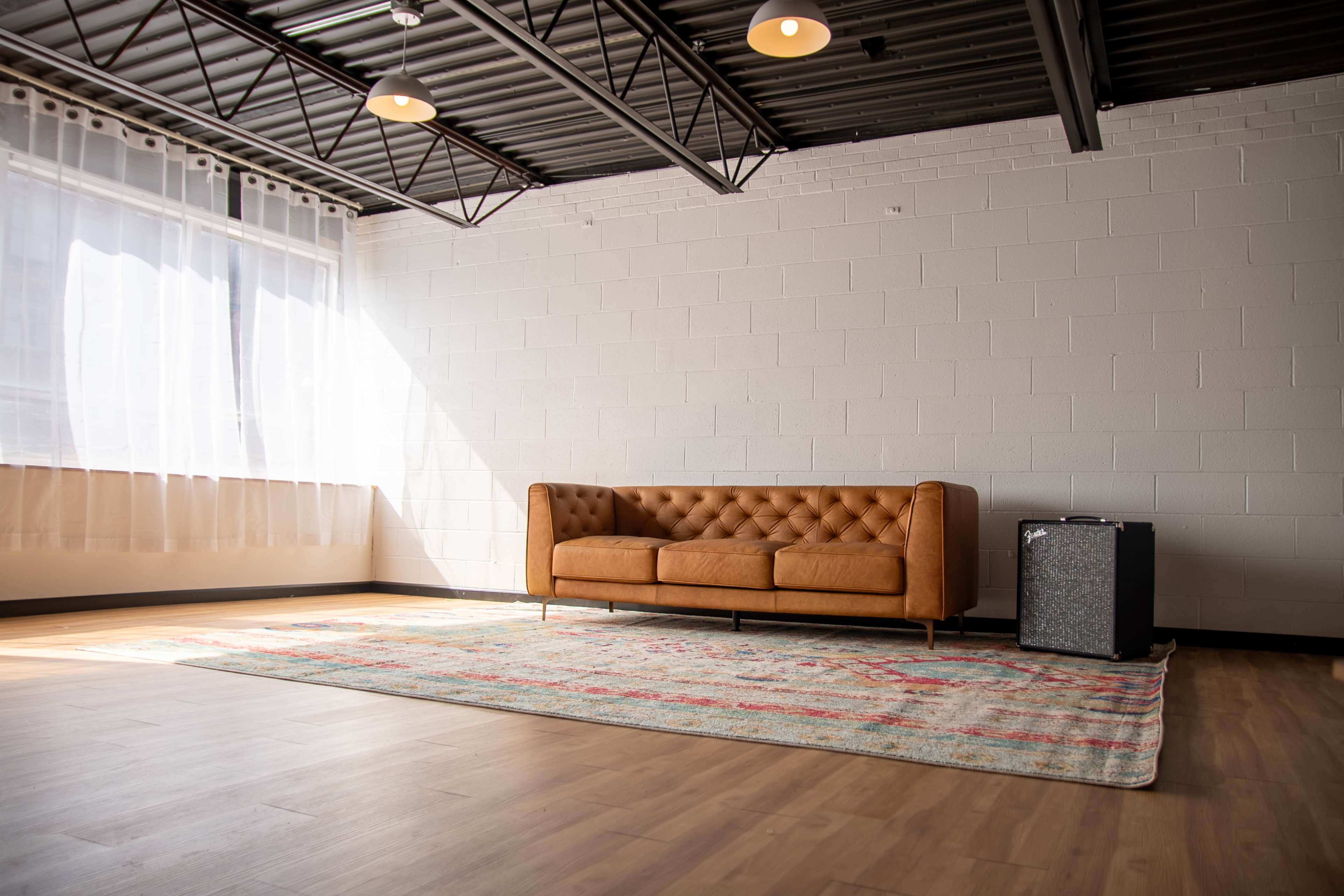 The image shows a well-lit room featuring a tufted brown leather sofa, a patterned rug, and an amplifier in the corner.