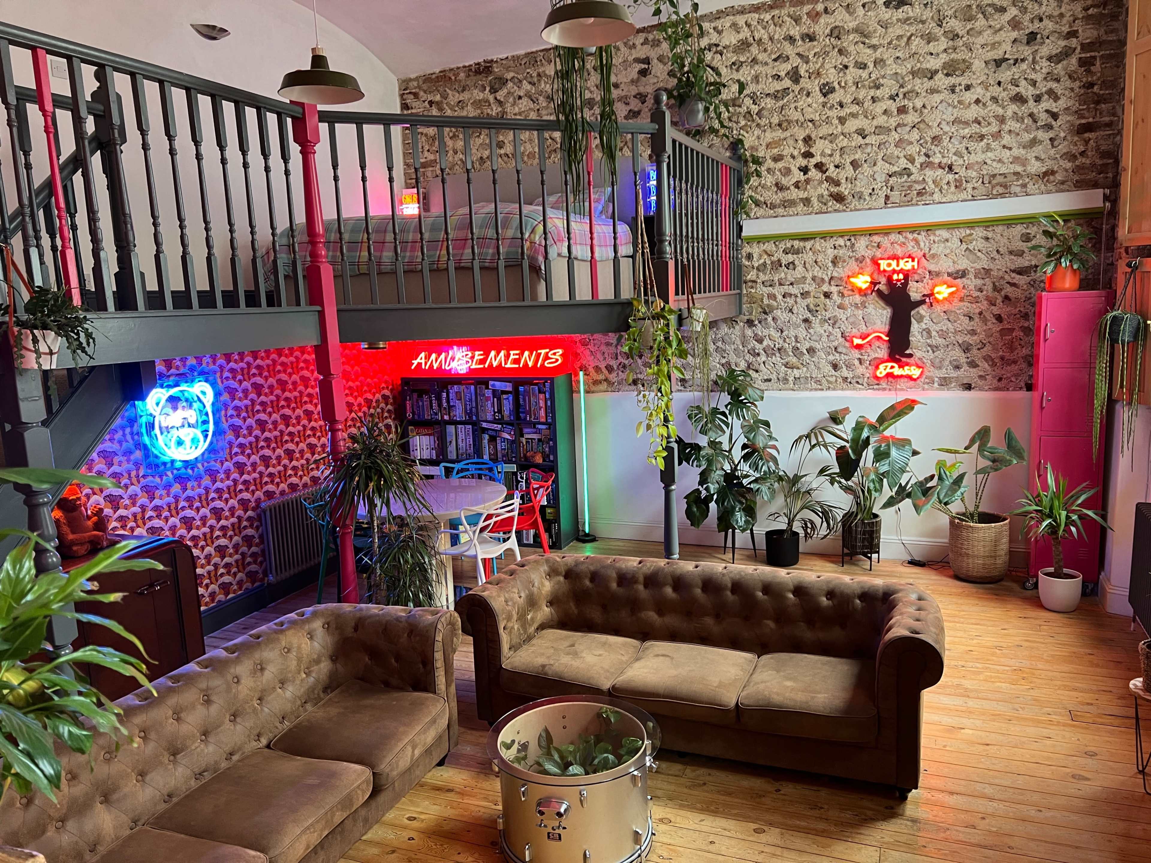 A spacious room featuring a loft area with a bed, two brown leather sofas, vibrant neon signs, and various indoor plants.