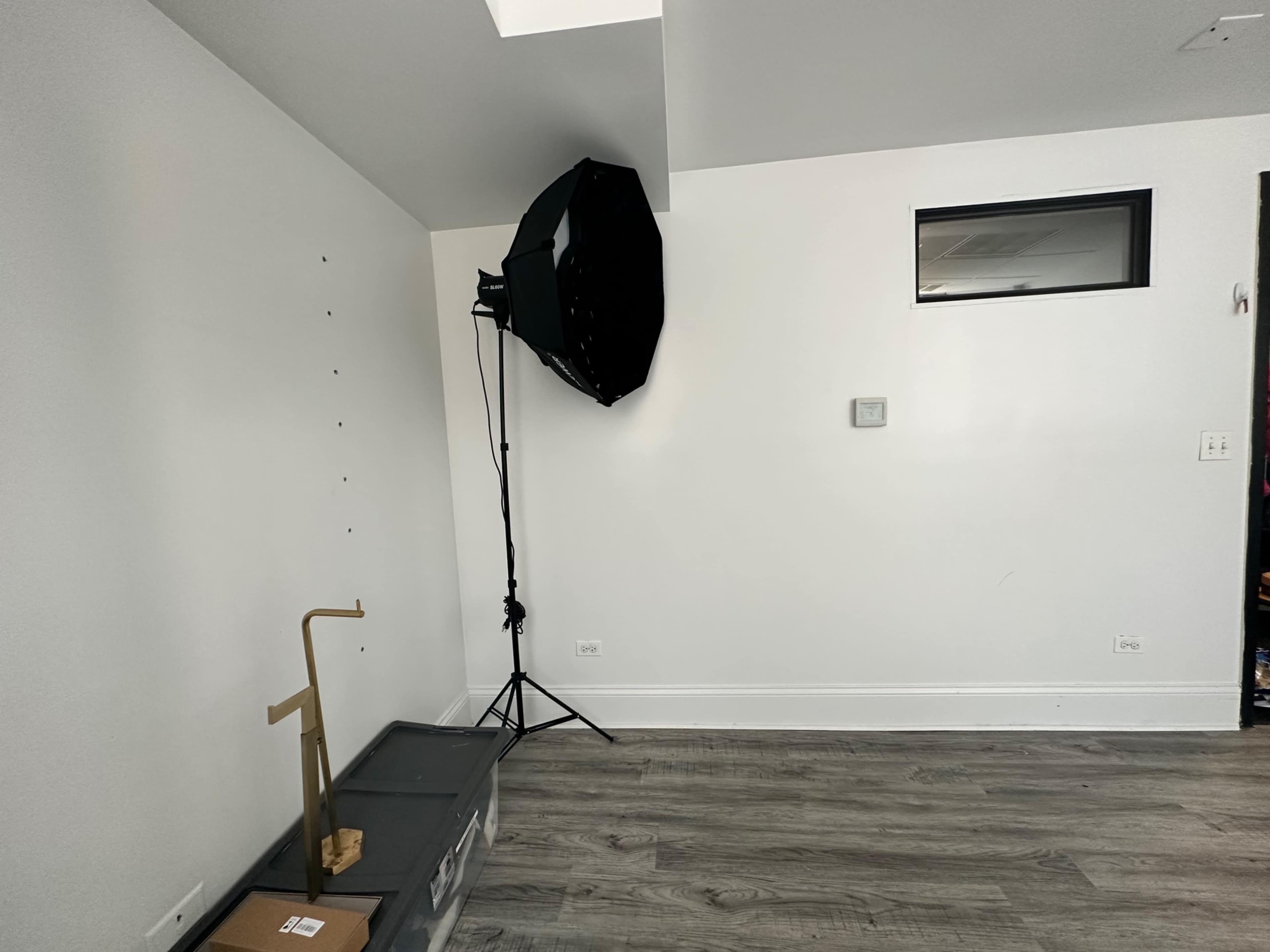 The image shows a minimalist room with a black softbox lighting setup in one corner and a storage box on the floor.