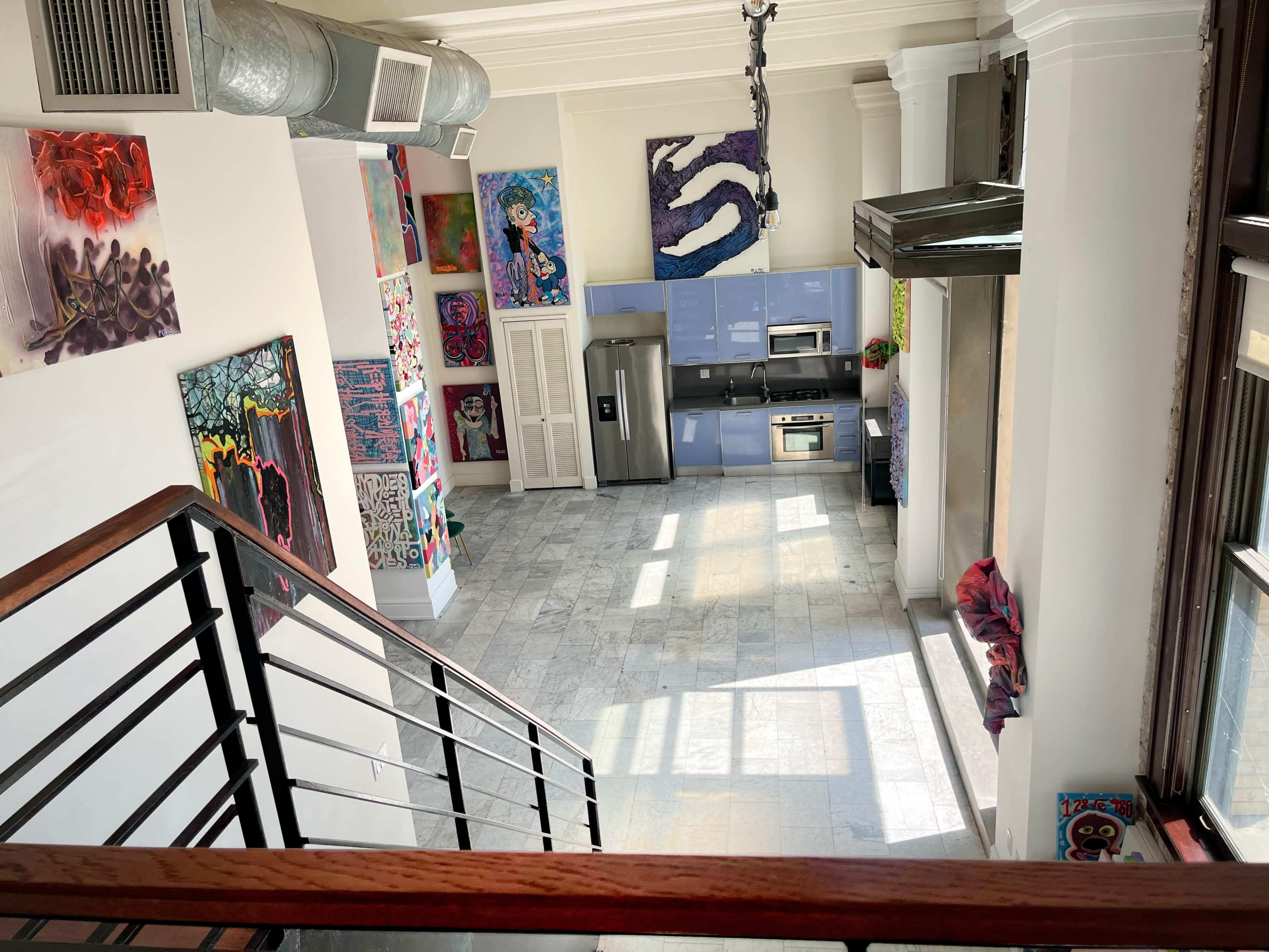The image shows a spacious kitchen and living area with modern appliances and colorful artwork on the walls, viewed from an upper level.