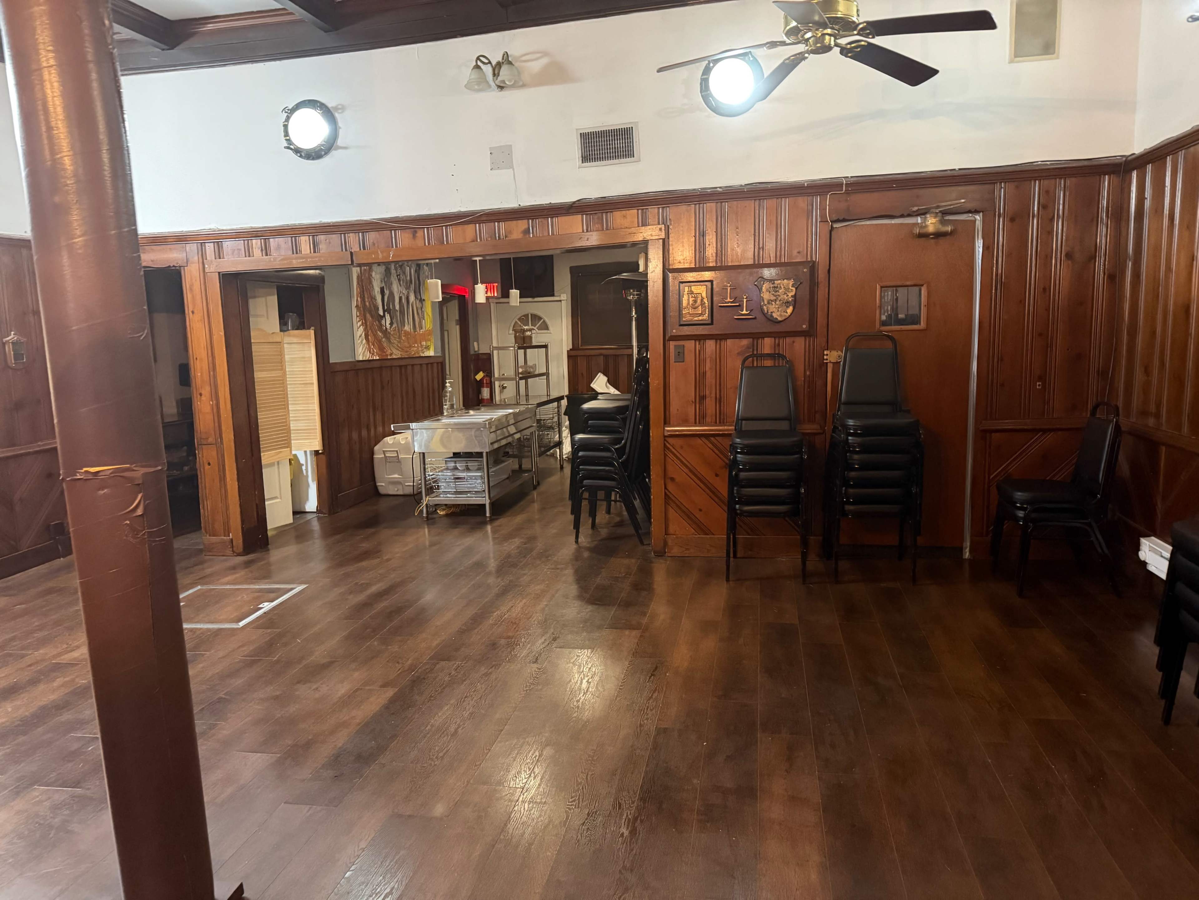 The image shows a wooden interior room with stacked chairs, polished floors, and two circular lights on the ceiling.