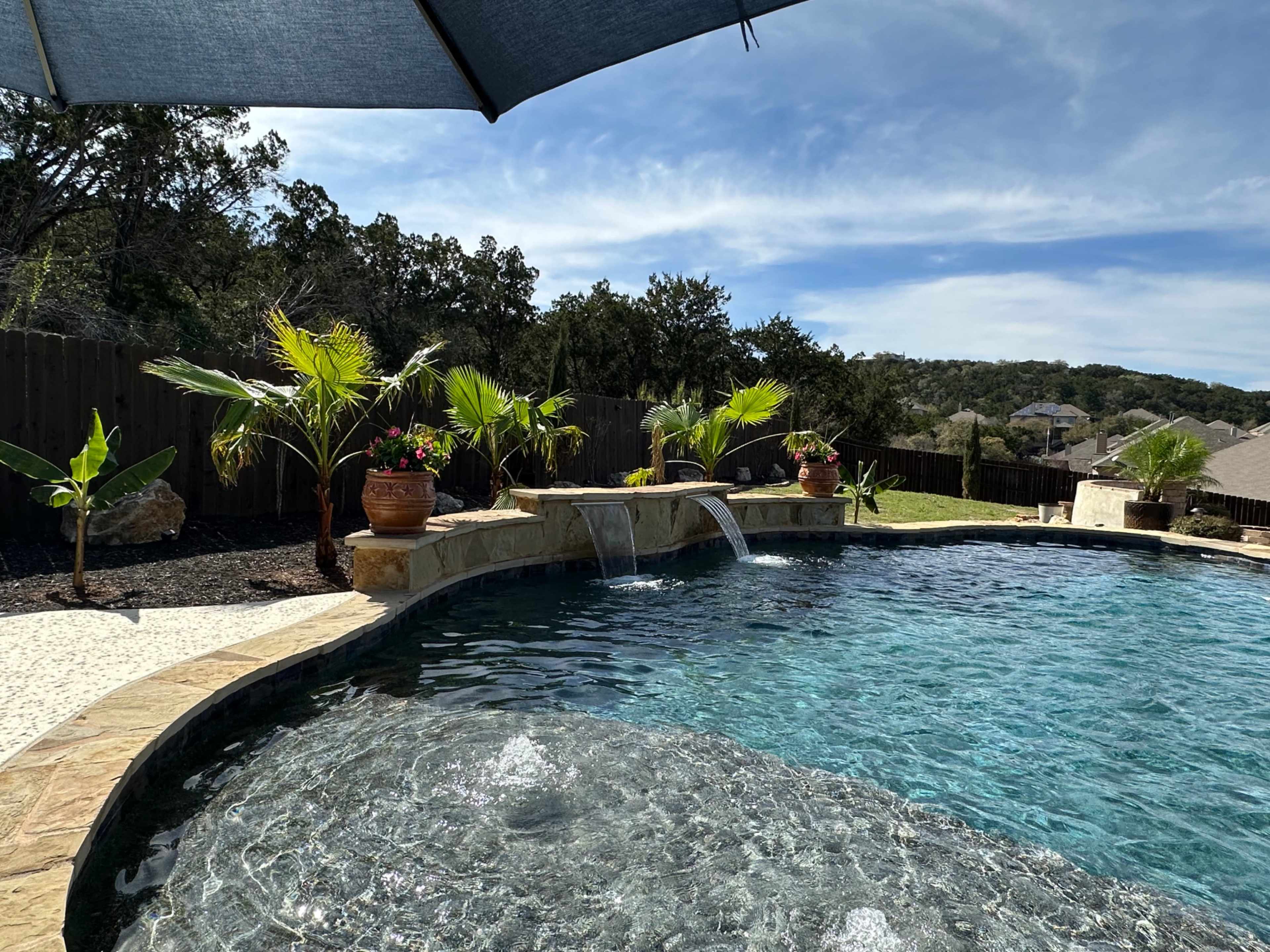 The image shows a swimming pool with a stone border, featuring waterfalls and potted plants, under a shaded area with a blue sky and trees in the background.