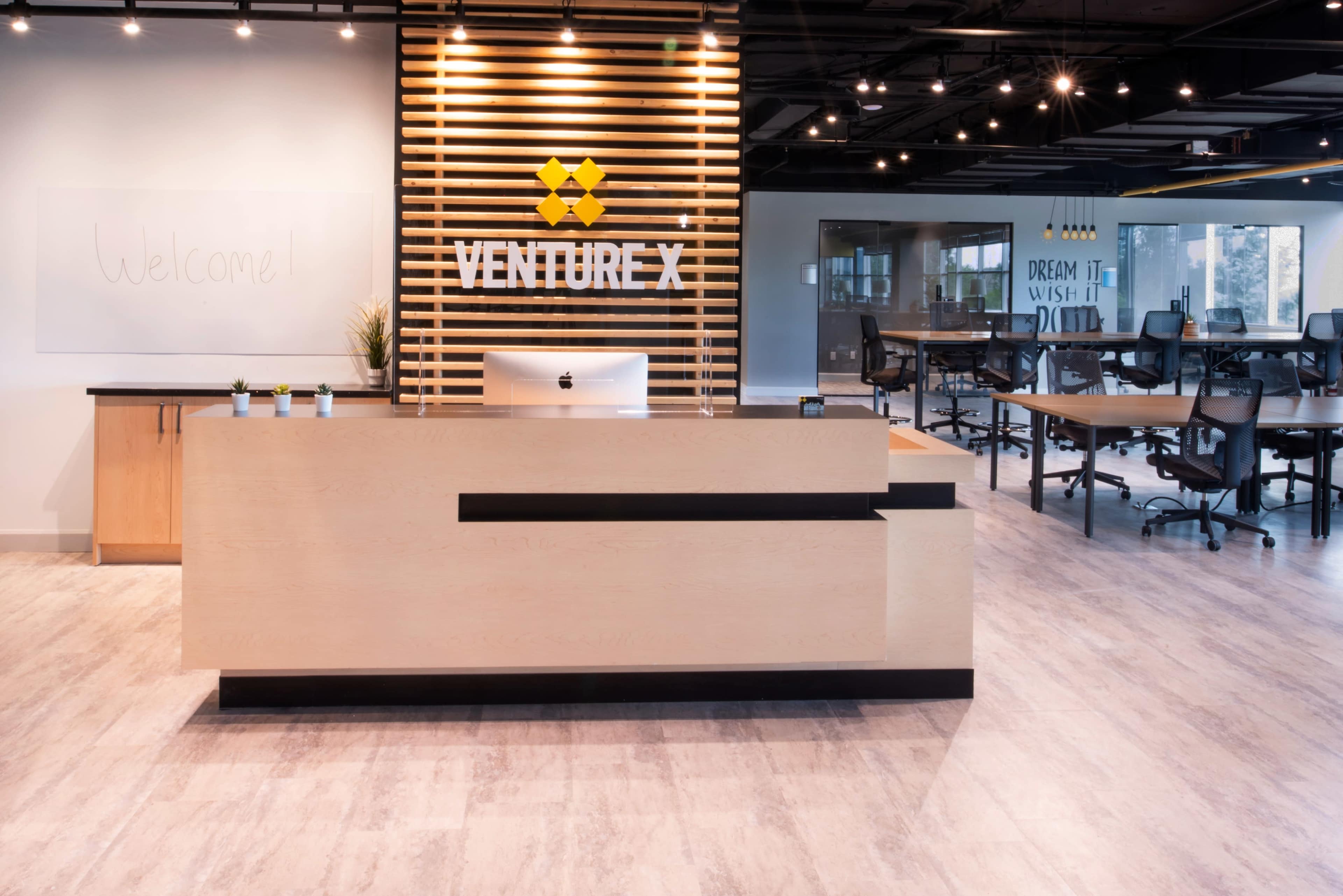 A modern reception area with a wooden front desk, a computer, and a wall featuring the Venture X logo, along with a welcoming message and a spacious work environment.