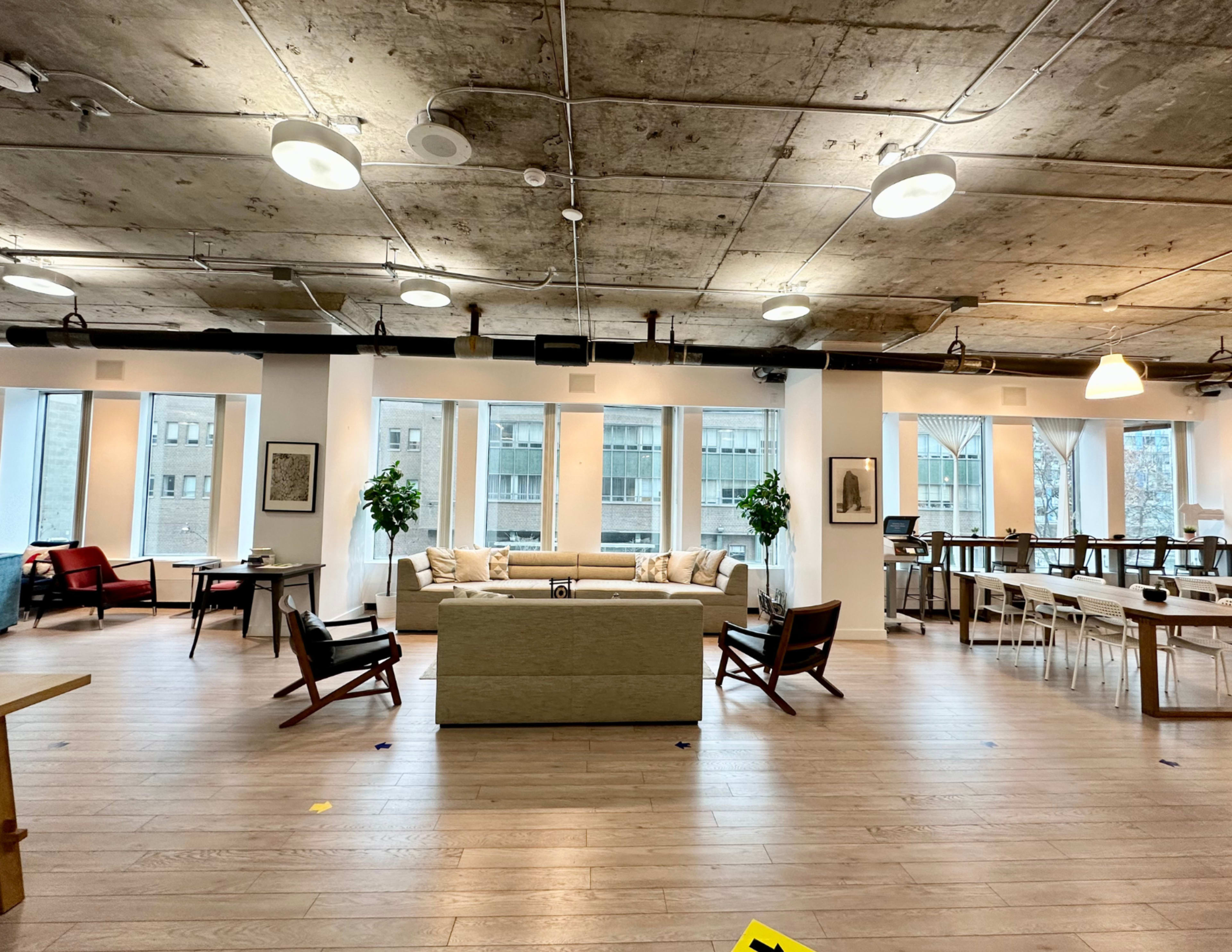 A spacious, modern office interior with a seating area featuring a light-colored sofa, several chairs, large windows, and wooden tables.