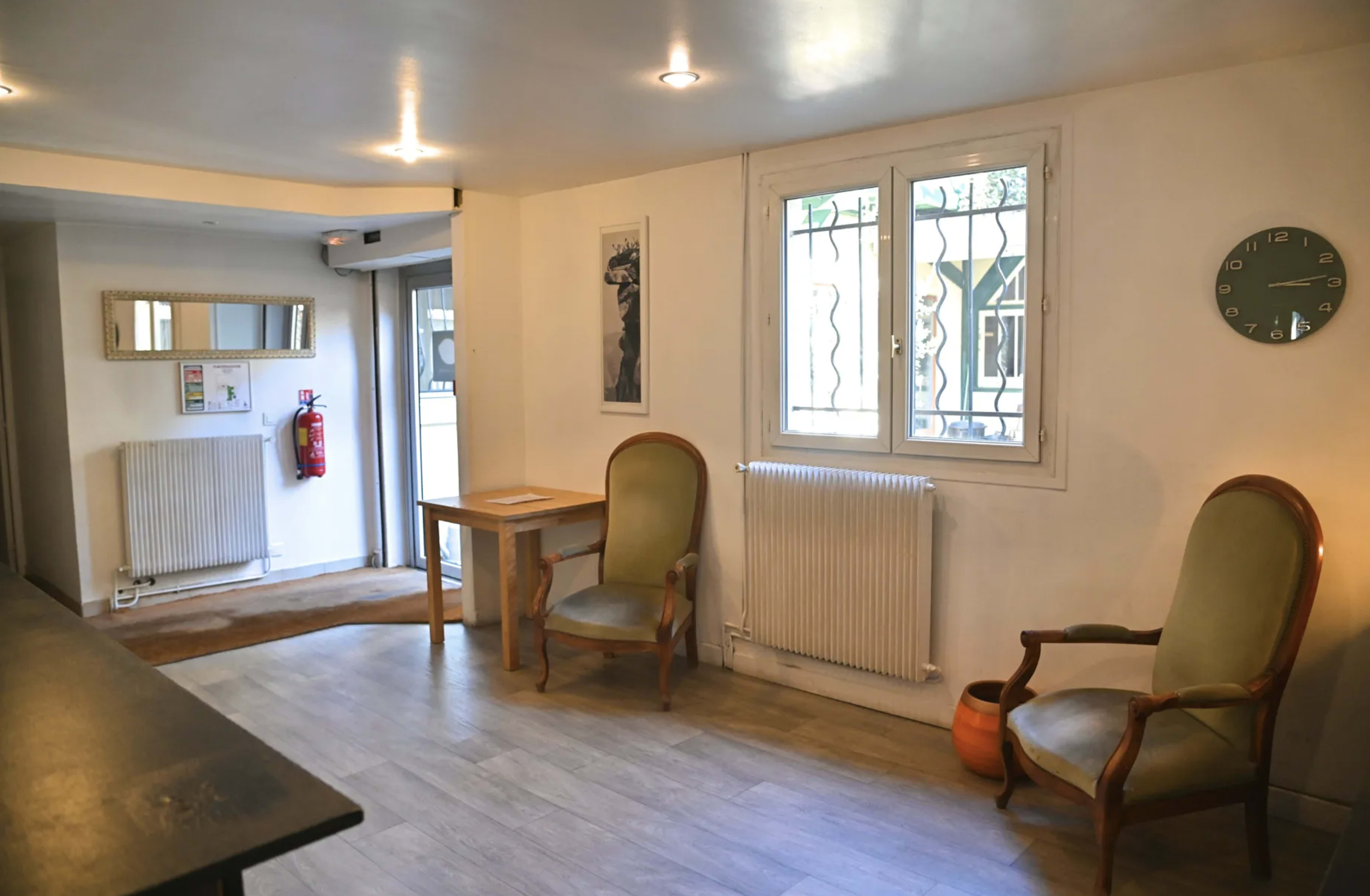 A simple interior of a room with two chairs, a table, a wall clock, a mirror, and a window with bars, leading to an exterior door.