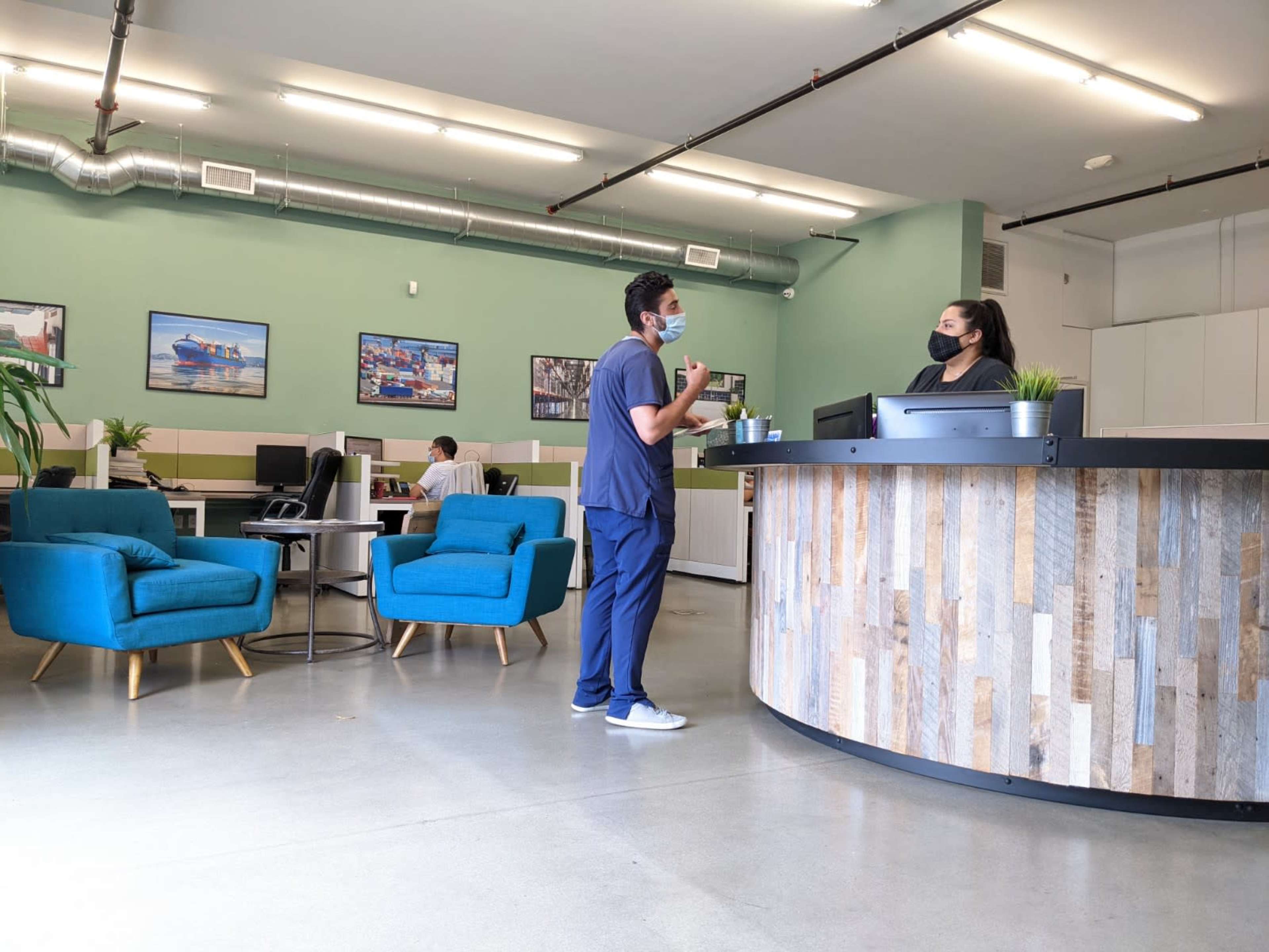 A reception area features two blue chairs, a front desk with two people interacting, and another individual seated at a workstation in the background.