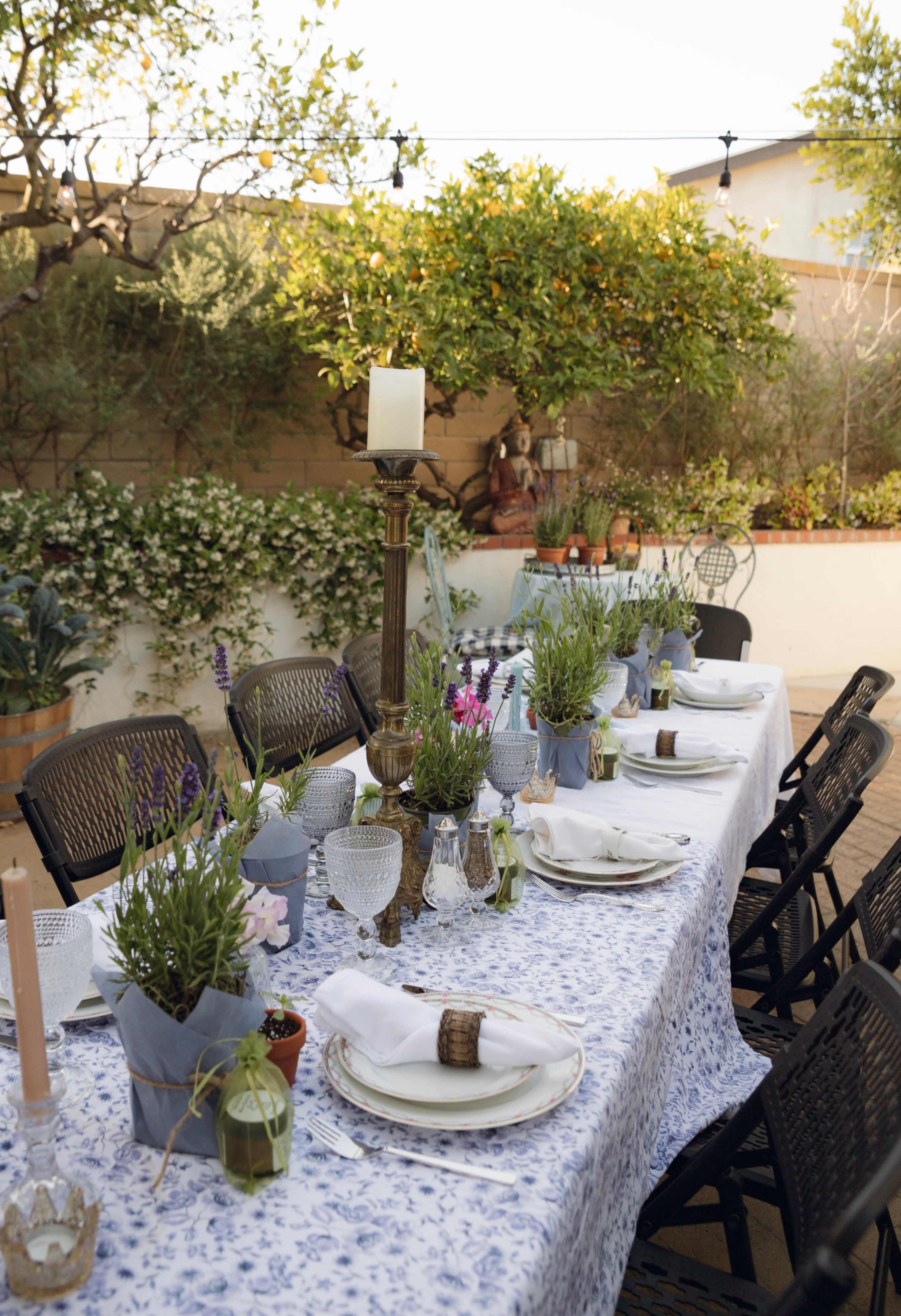 A long outdoor dining table is set with floral tablecloths, potted plants, candles, and elegant tableware, surrounded by greenery and decorative lighting.