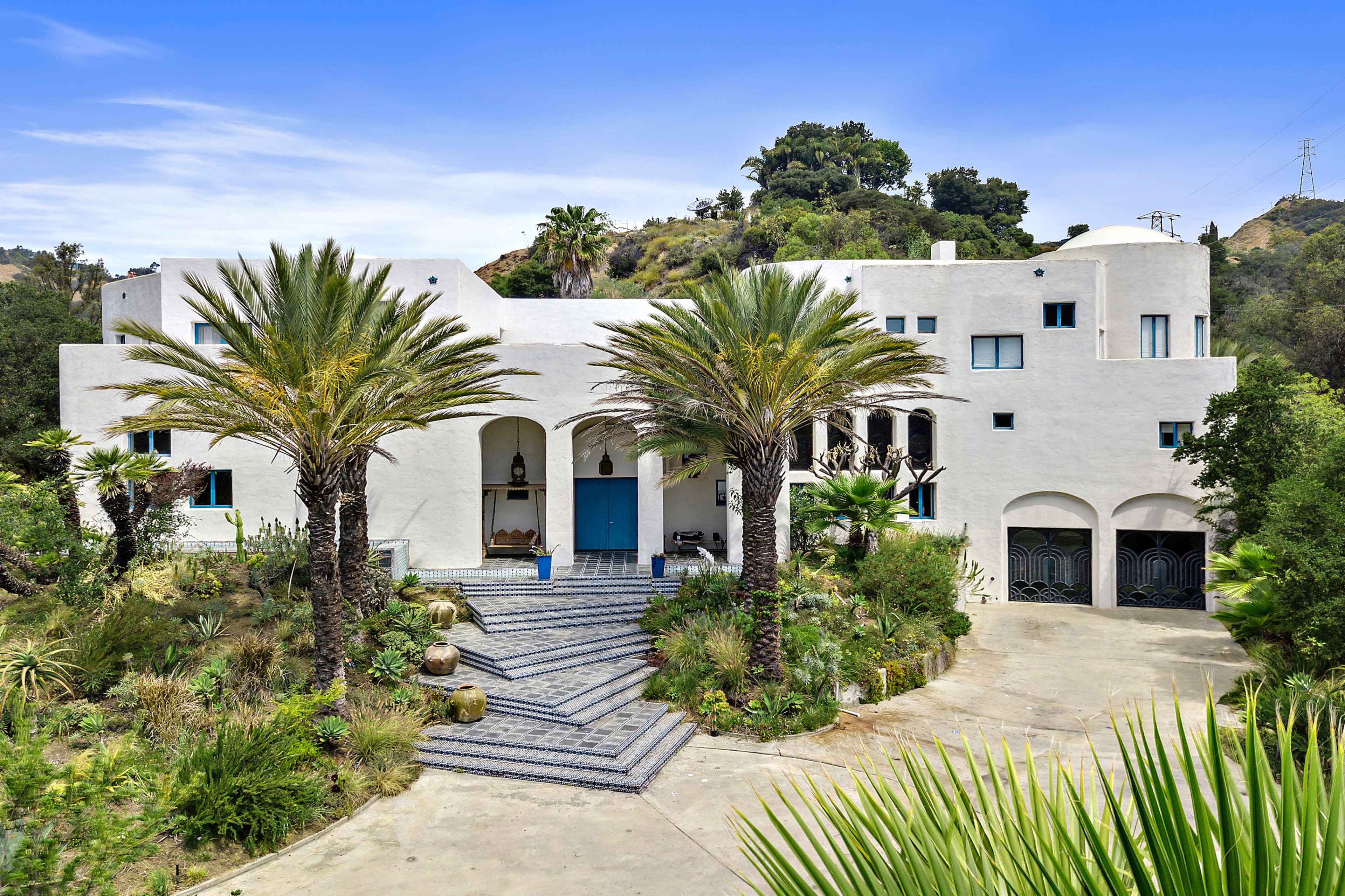 The image shows a large white house with a blue front door, surrounded by palm trees and tropical landscaping.