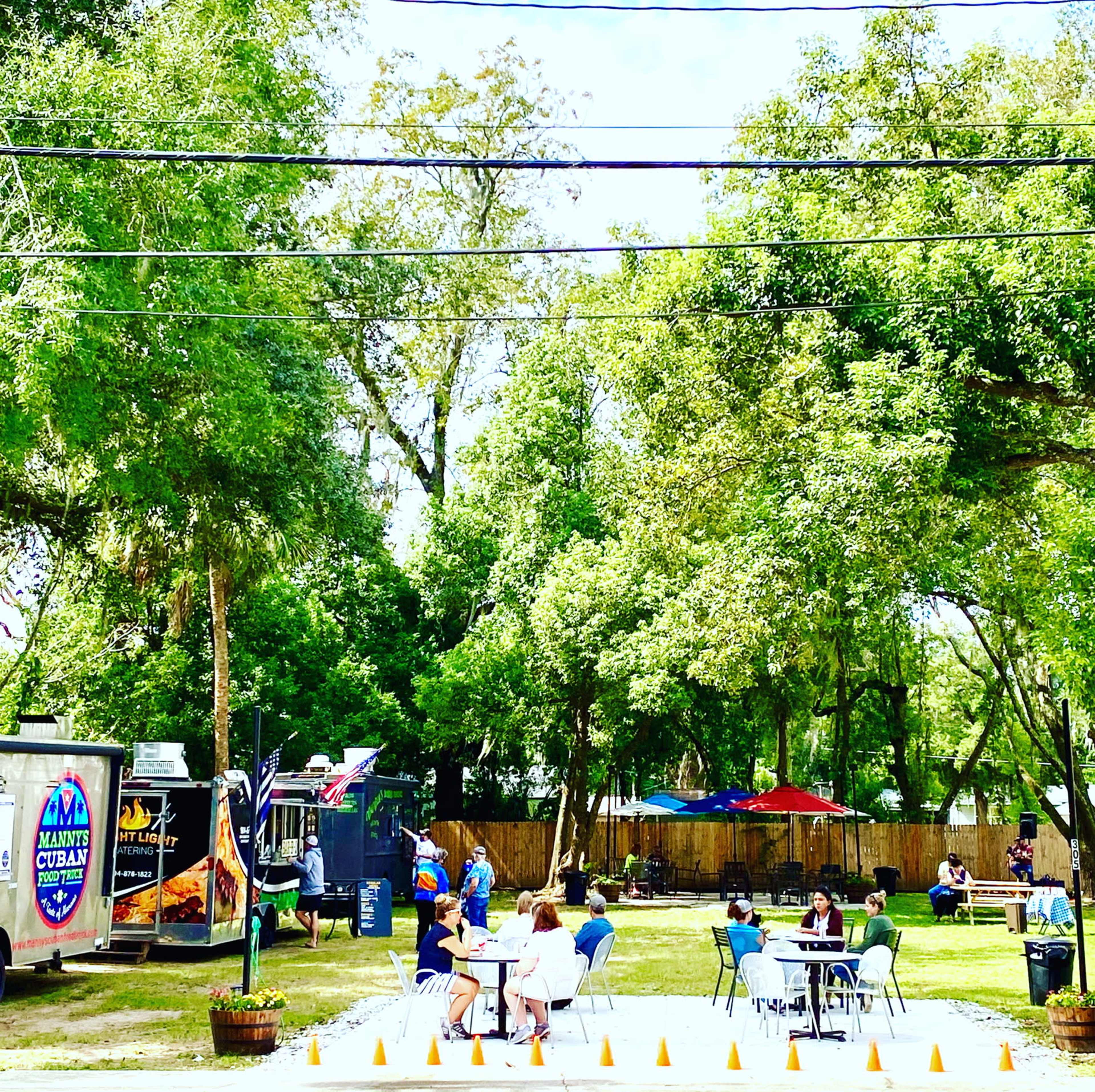 The image shows a vibrant outdoor food truck area with several patrons dining at tables under tree shade.