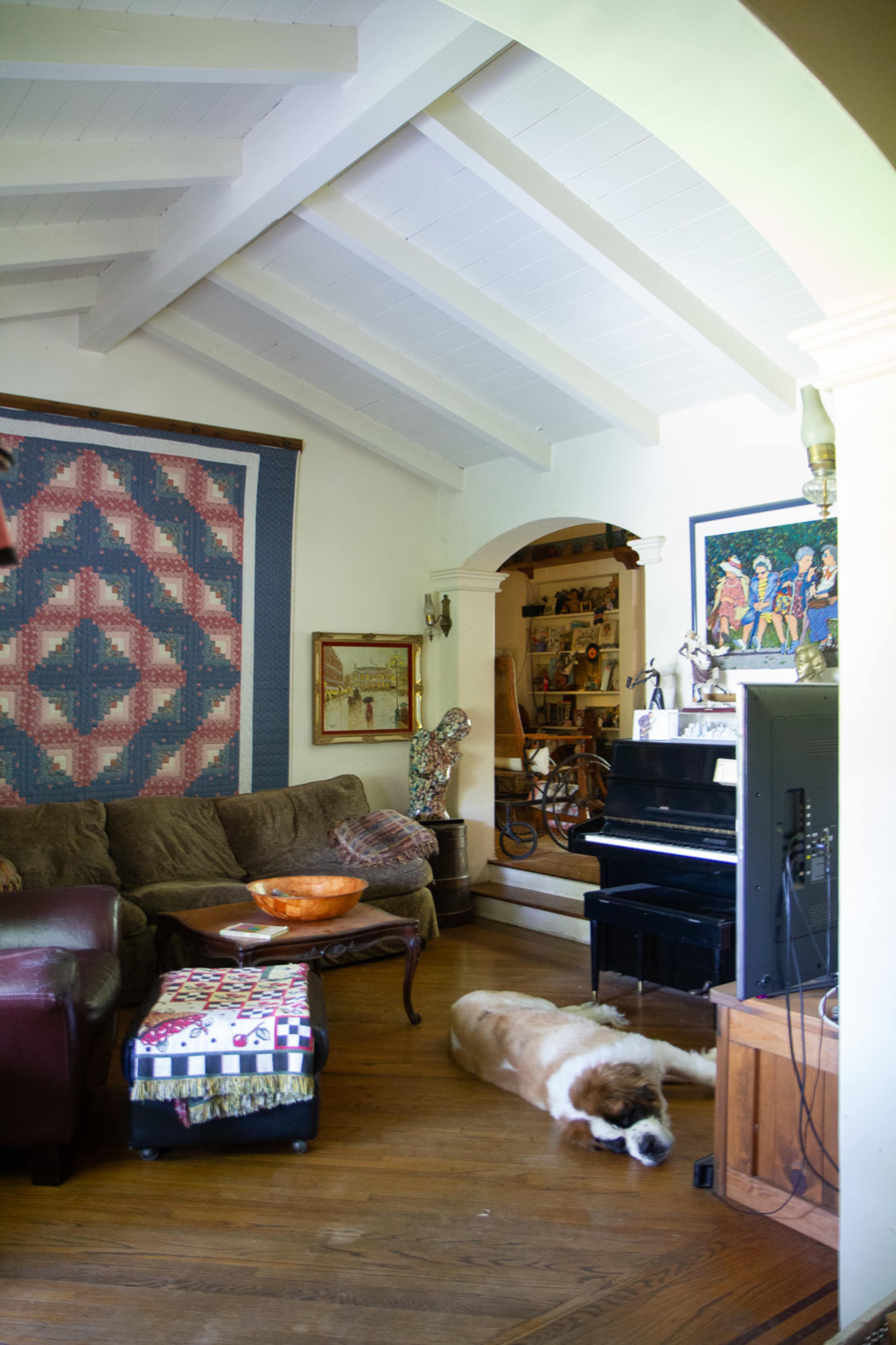 A cozy living room features a large couch, a quilted wall hanging, a piano, and a Saint Bernard dog lying on the wooden floor.