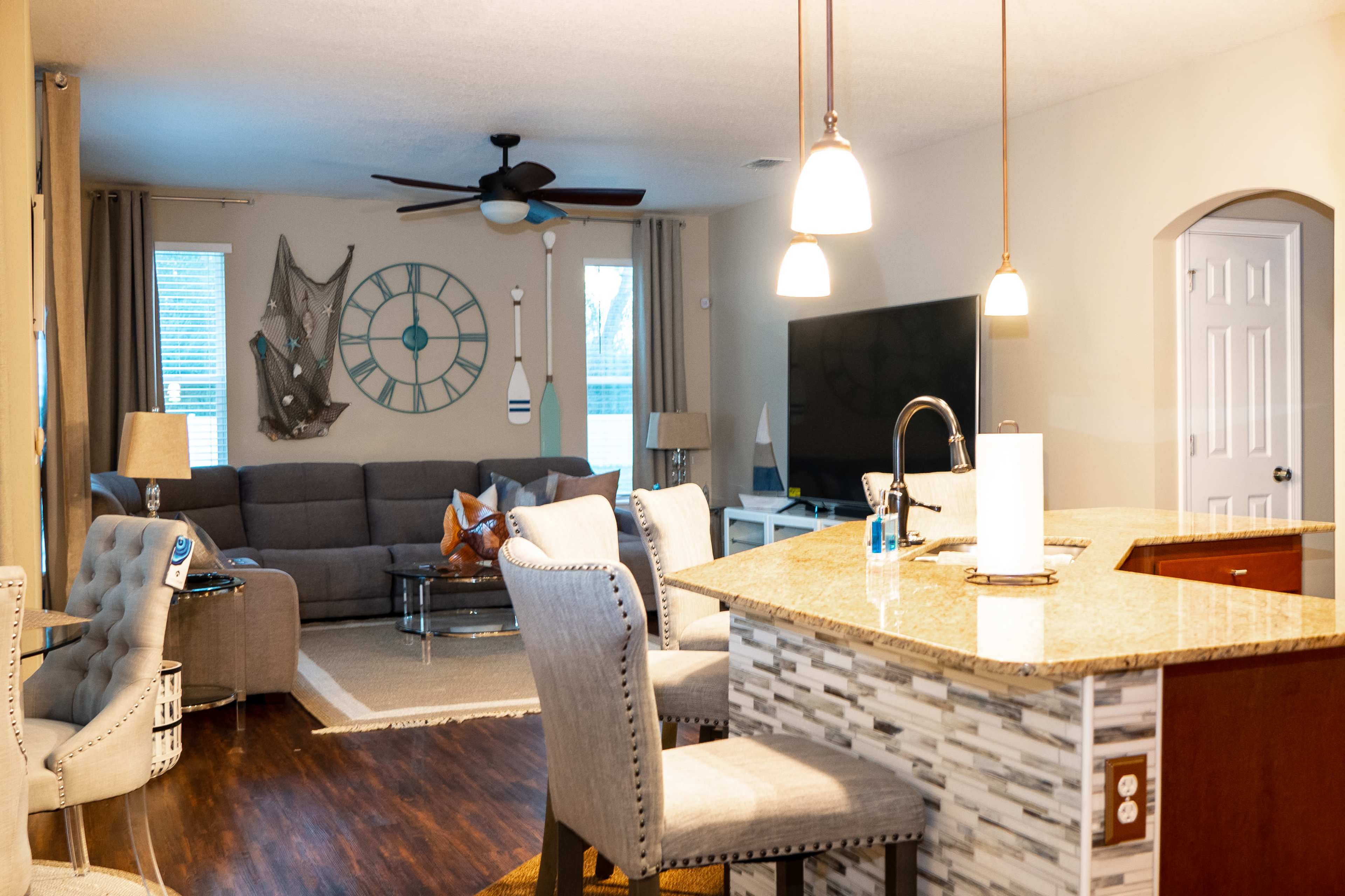 A modern living room with a gray sectional sofa, a large wall clock, and a kitchen bar with stools, featuring wooden flooring and neutral-colored walls.