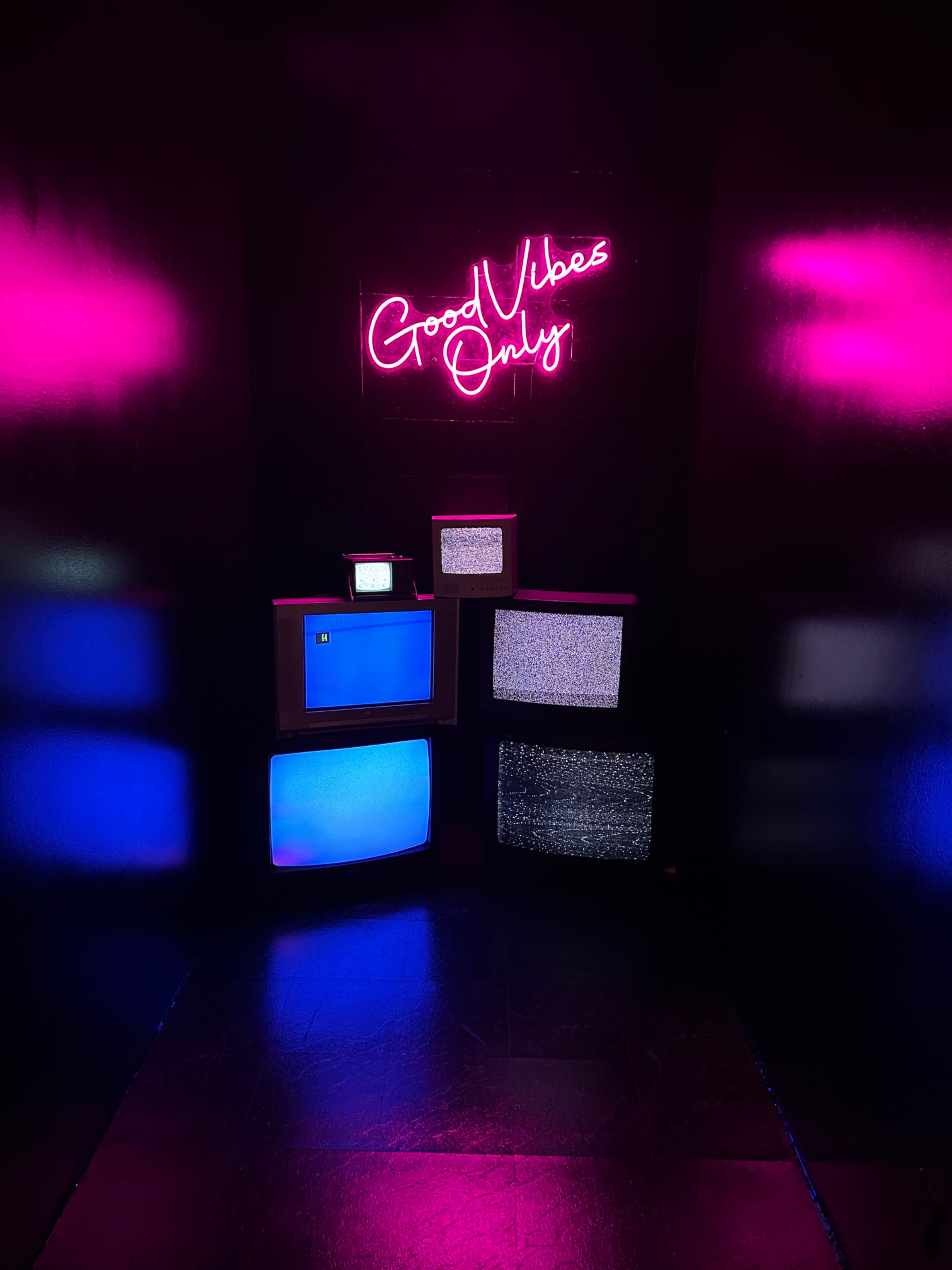 A collection of retro televisions is arranged in a dark room, illuminated by a neon sign that reads "Good Vibes Only."